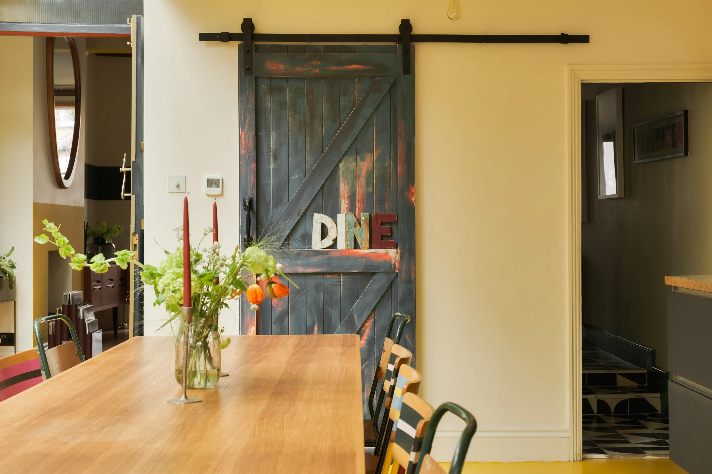 Dining area with a wooden table and colorful chairs, a glass vase with flowers, red candles, and a painted sliding barn door with the word "DINE" on it.