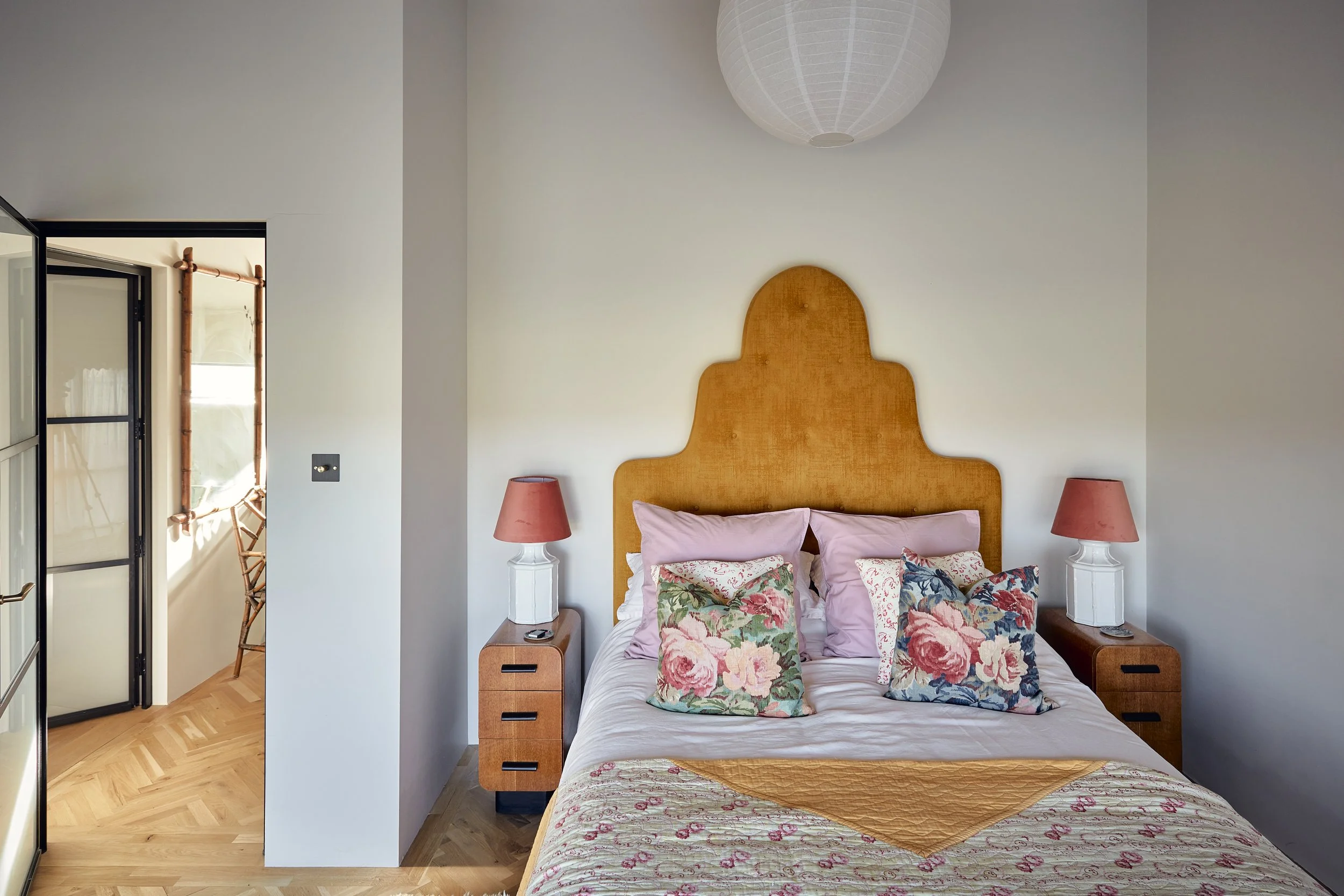 A bedroom with a bed featuring a wooden headboard, pink and floral pillows, and a patterned blanket. Two matching pink lamps are on white bedside tables, and there's a paper lantern ceiling light. An open doorway leads to a balcony with a chair and c
