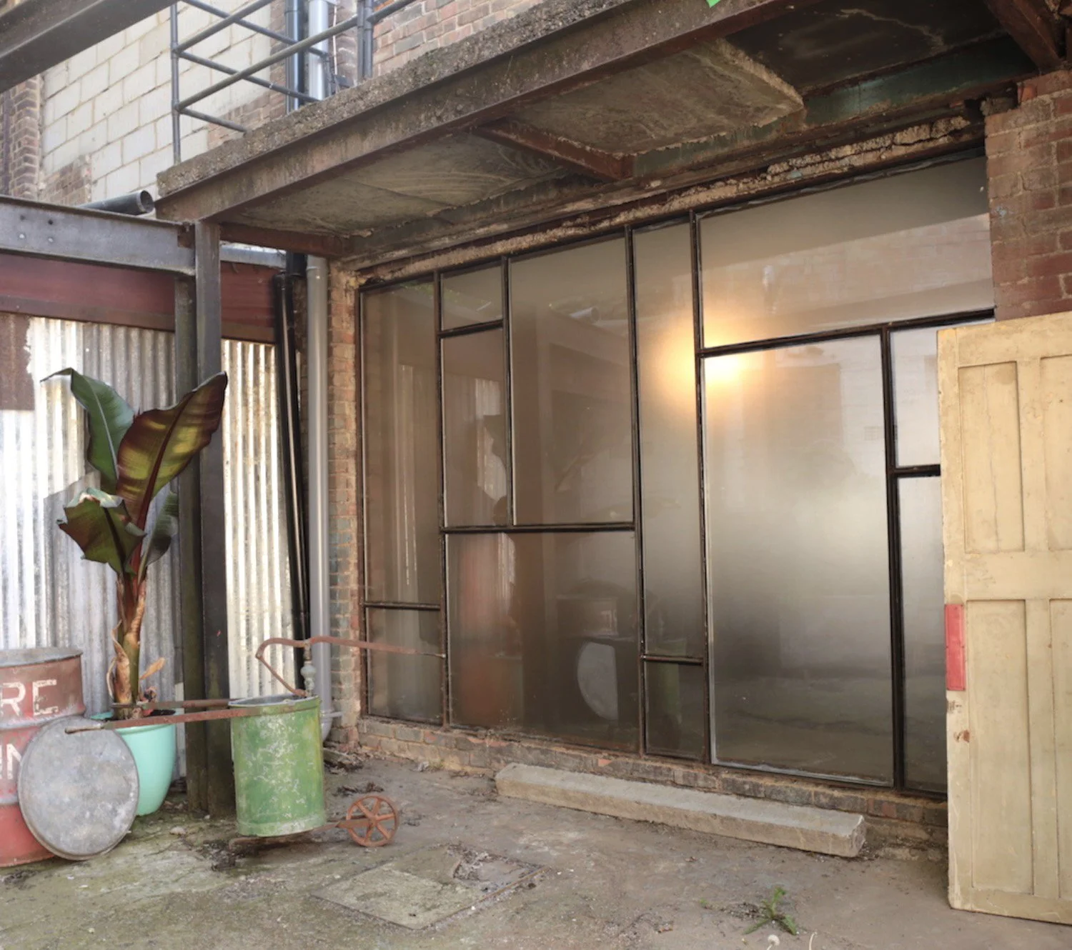 An industrial-style brick building exterior with a large glass and metal window or door frame, construction materials, a potted plant, and some weathered containers and tools in the foreground.