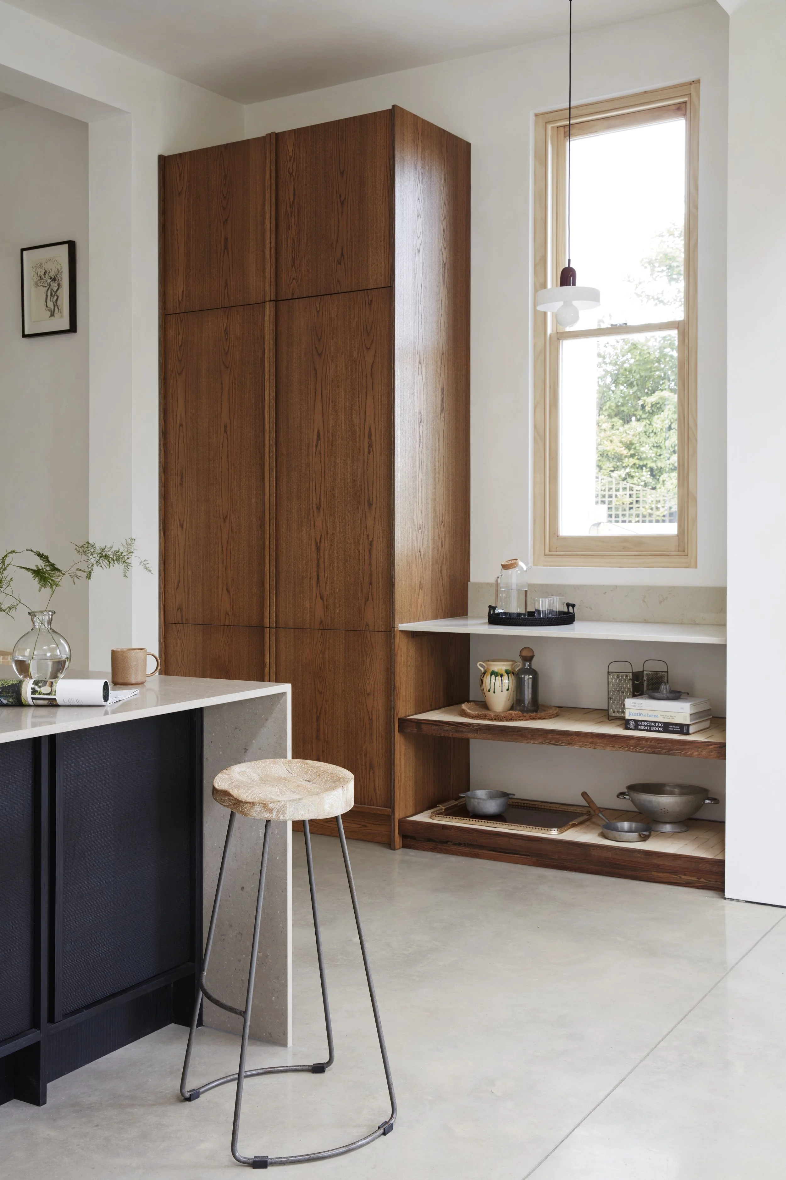 A modern kitchen corner featuring a wooden cabinet, a window with a wooden frame, a hanging light fixture, a small wooden shelf with decorative items, a black and concrete island, and a wooden stool.