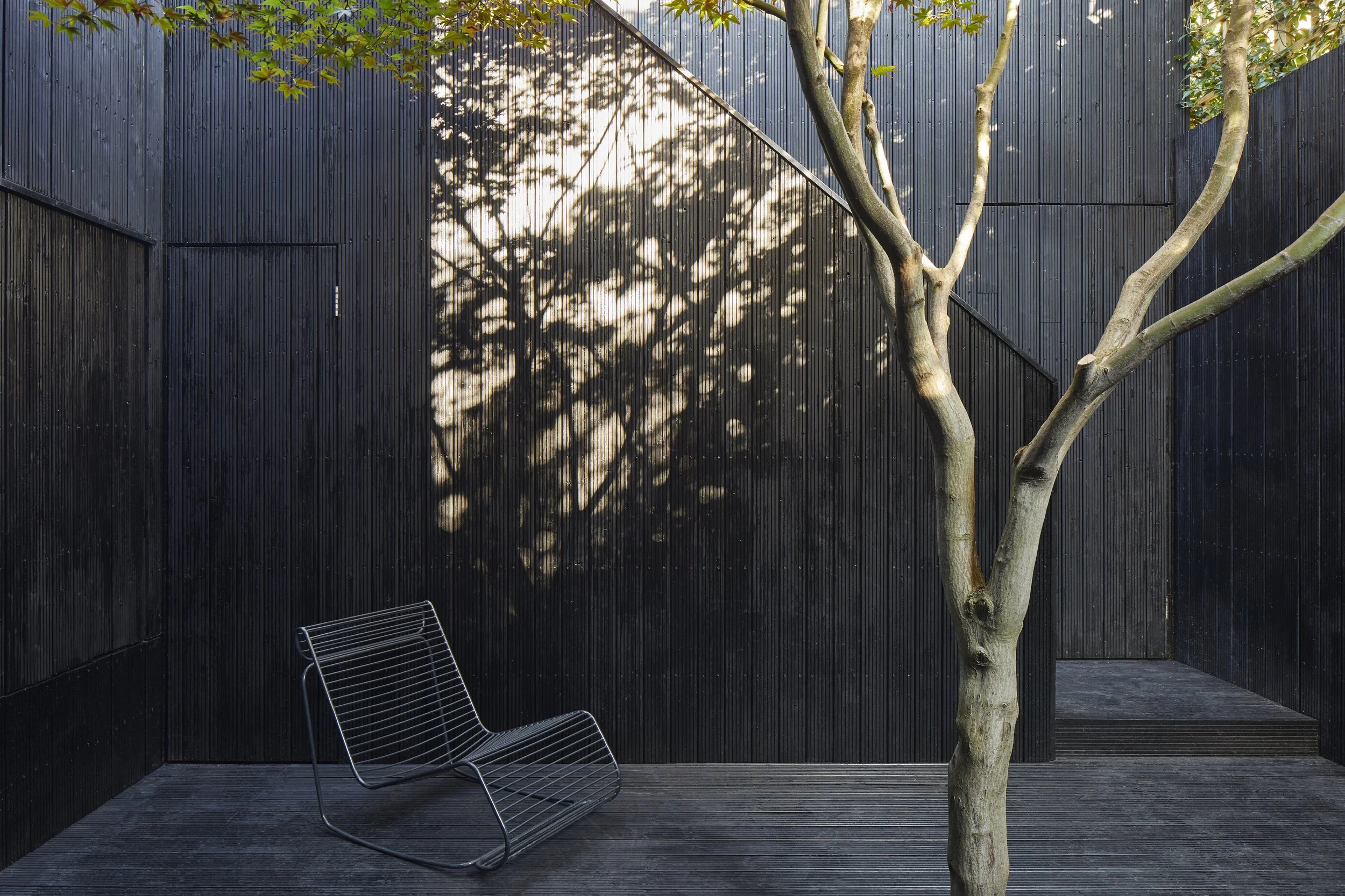 A modern outdoor patio with black wooden walls, a metal chair, a tree with shadows cast on the wall, and wooden flooring.