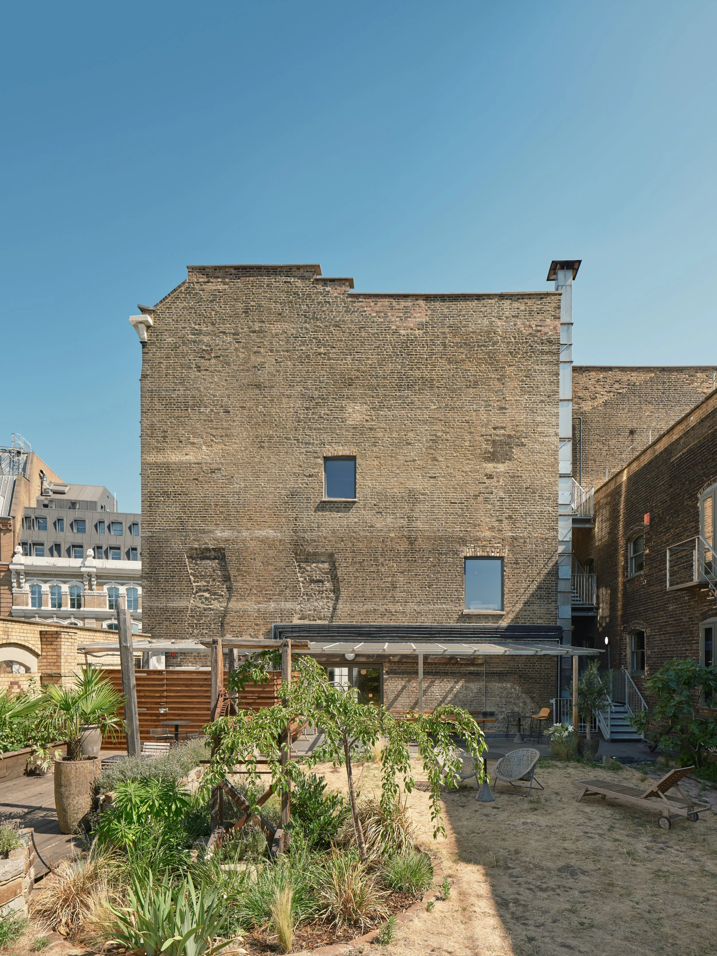Urban backyard with garden plants, lounge chairs, and patio furniture, with a large brick building in the background under a clear blue sky.
