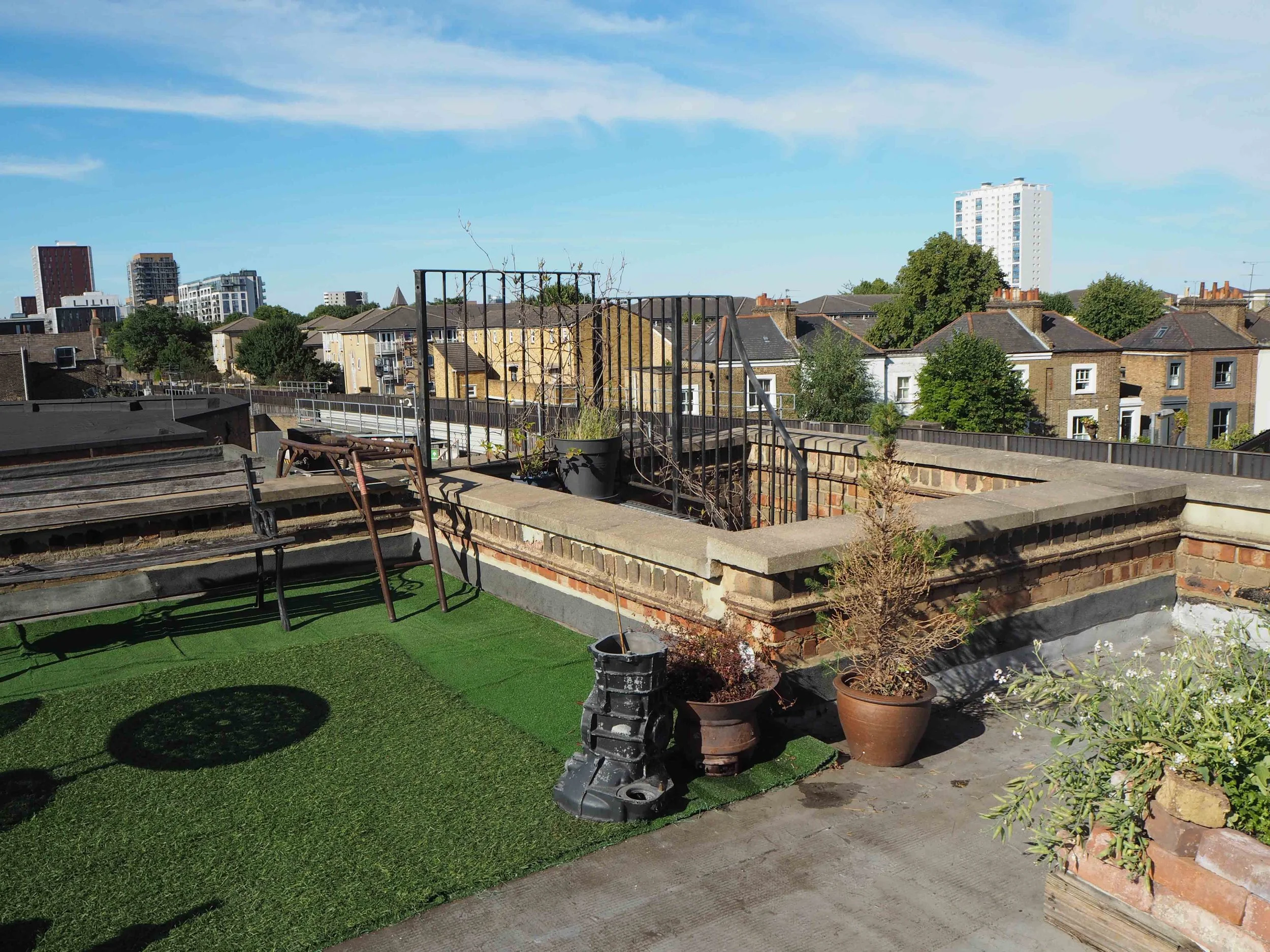 Rooftop with artificial grass, potted plants, and outdoor furniture, overlooking a cityscape with residential houses and tall buildings.