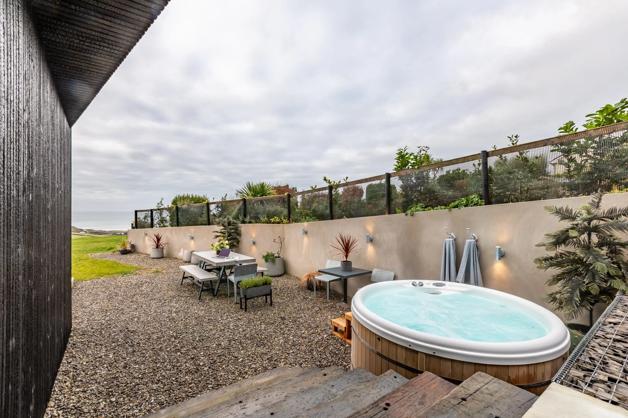Outdoor patio with hot tub, tables, and chairs, surrounded by potted plants, on a gravel surface with a view of the ocean in the background.