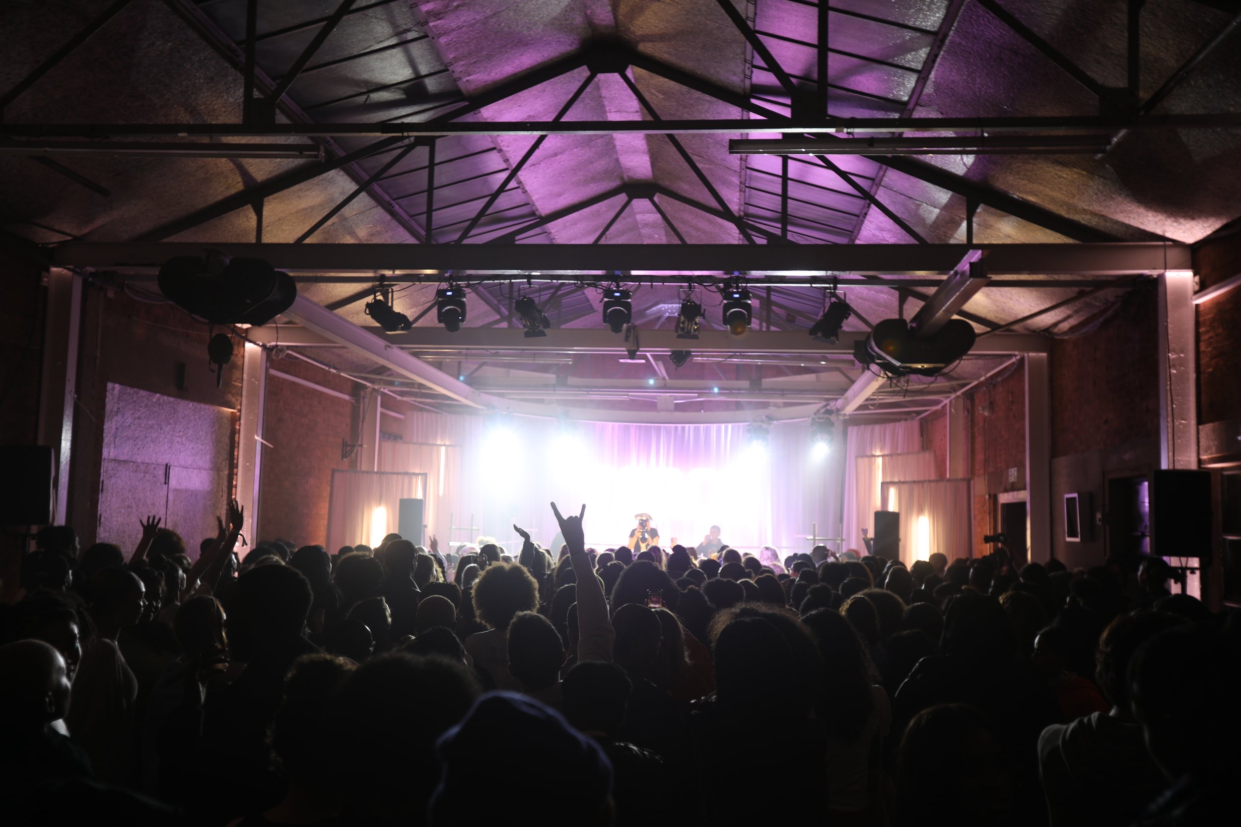 A large crowd watching a live concert or performance on stage with bright lights and purple lighting effects in an indoor venue.