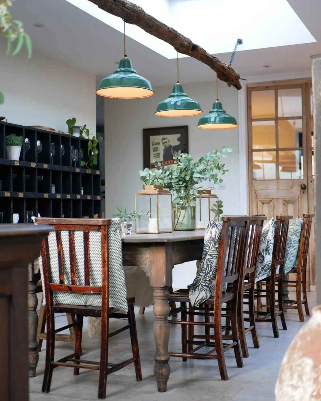 A cozy dining room featuring a round wooden table with green pendant lights hanging above, a large vase with greenery as a centerpiece, and a set of wooden chairs with eclectic cushions. In the background, a black wall-mounted shelving unit holds pot