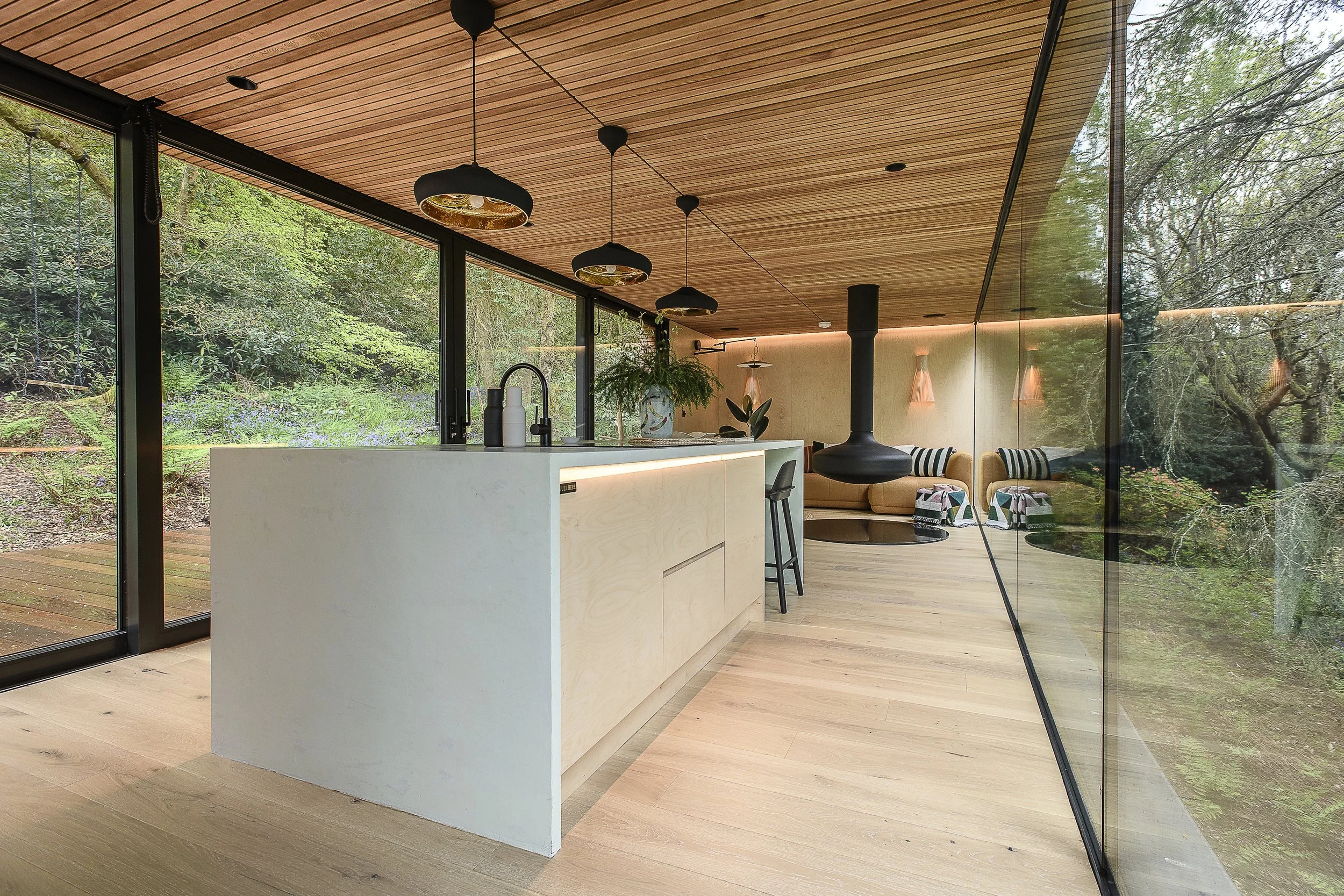 Modern open-concept living space with a kitchen and seating area, featuring large glass walls offering a view of a lush forest outside. The interior has a wooden ceiling, a white kitchen island, pendant lights, and neutral-colored furniture.