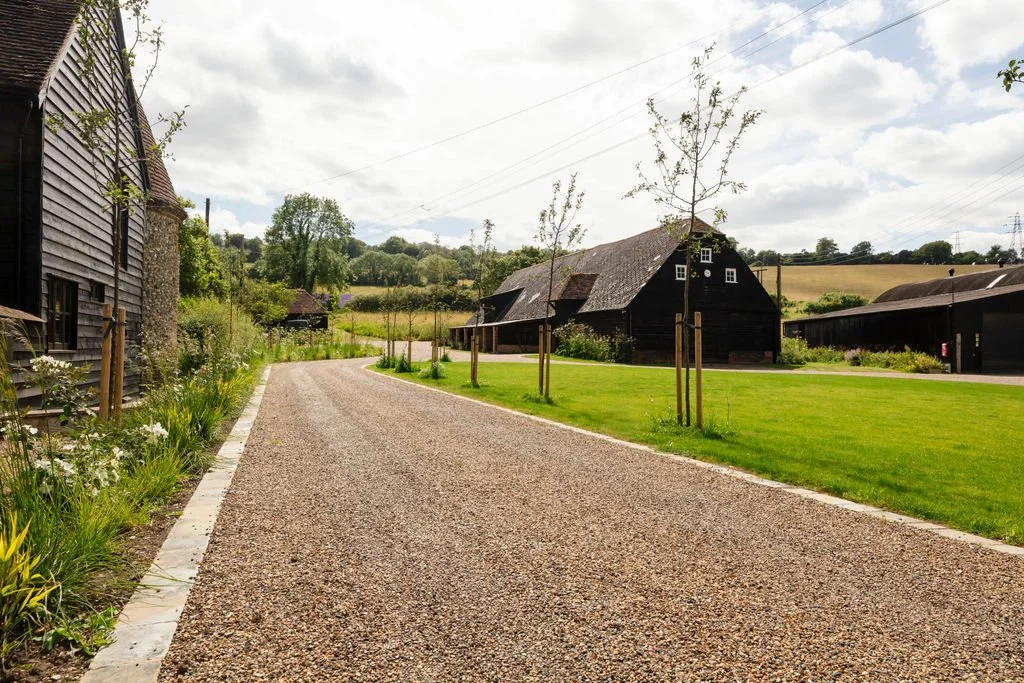A rural farm scene with a gravel pathway, wooden barns, and small trees lining the path, surrounded by fields and greenery under a partly cloudy sky.