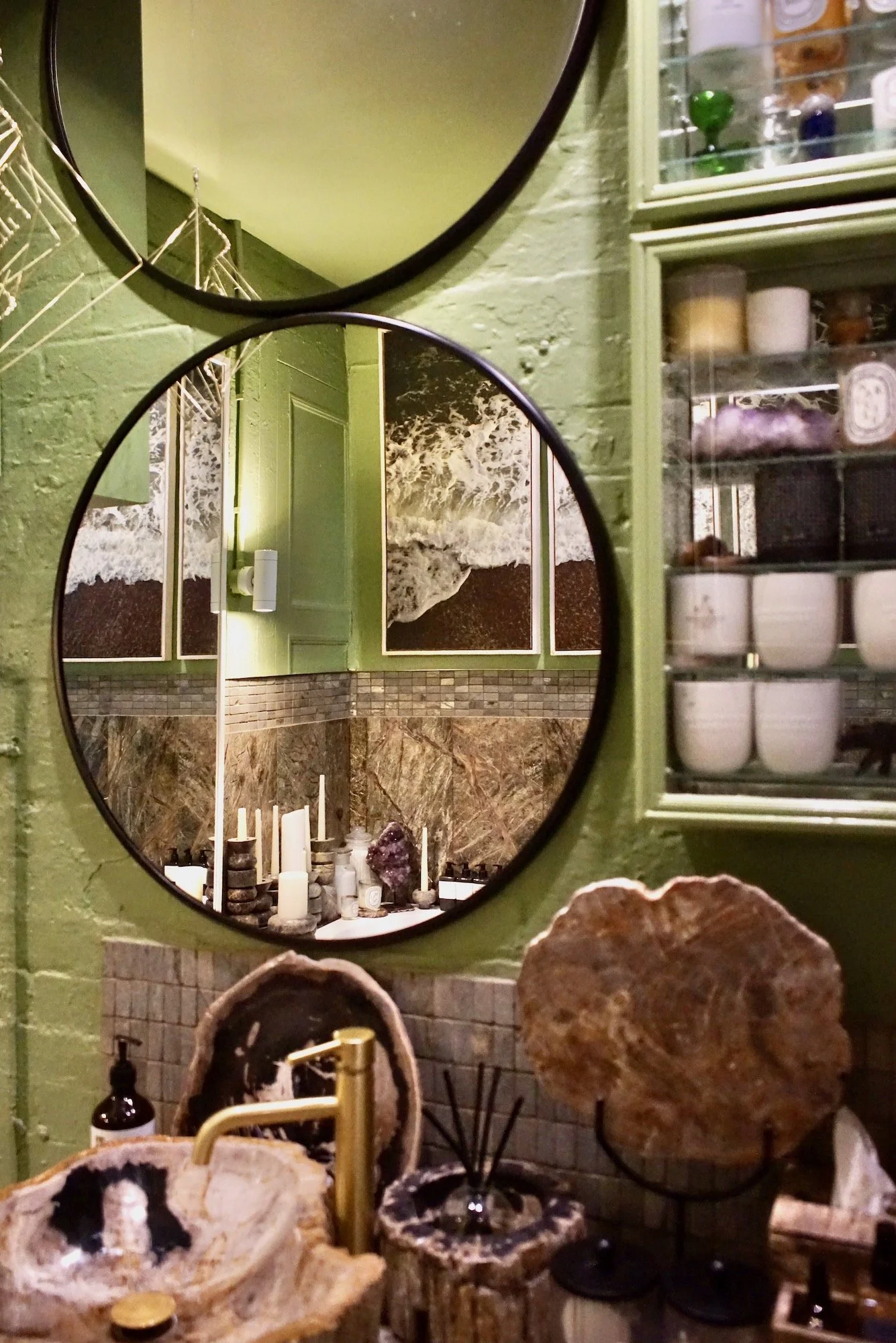 A corner of a room with two round mirrors on a green painted wall. The mirror reflects a decorated wall with ocean wave artwork, candles, and decorative items. To the right, there's a glass-fronted cabinet with shelves holding various small container
