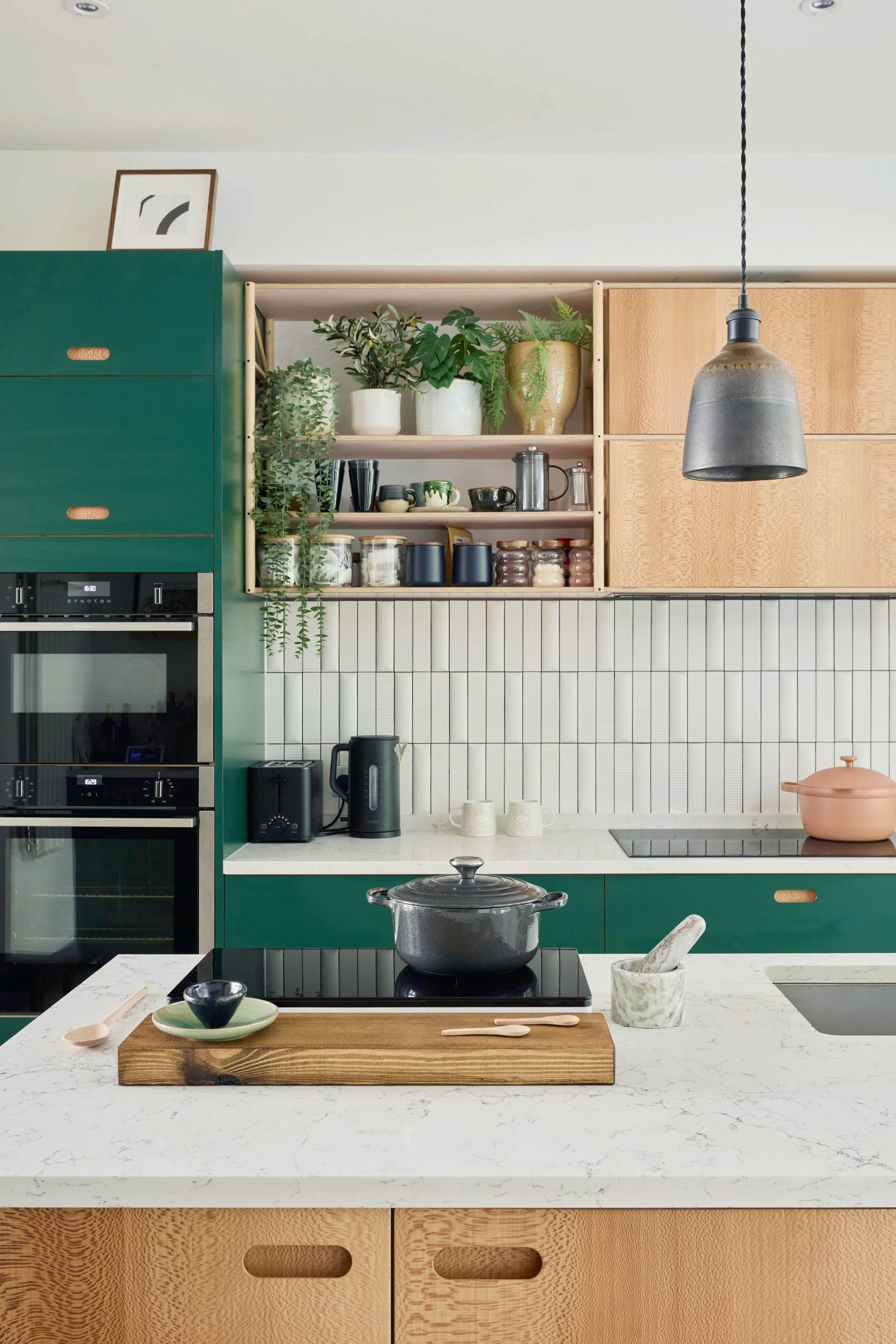 Modern kitchen with green cabinetry, open wooden shelves with plants and jars, marble countertop, black stovetop with pot, and pendant light.