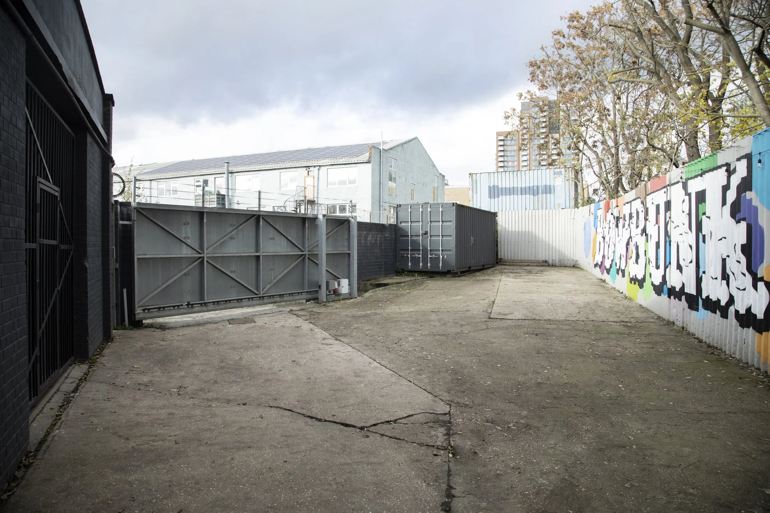 Empty concrete alley with graffiti-covered wall on the right and a gray metal gate on the left. Behind are industrial buildings and trees, with a cloudy sky overhead.