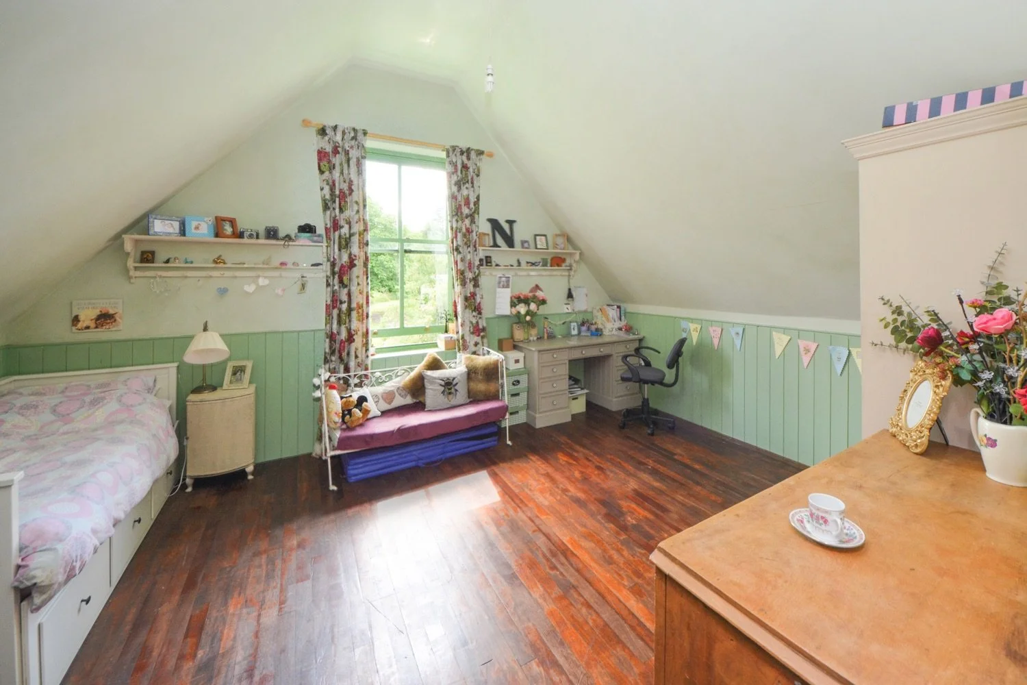 A cozy bedroom with a sloped ceiling, featuring a bed with pink bedding, a nightstand with a lamp, a window with floral curtains, a small sofa with pillows, a desk with a chair, and a dresser with flowers and decorative items, all on dark wooden floo