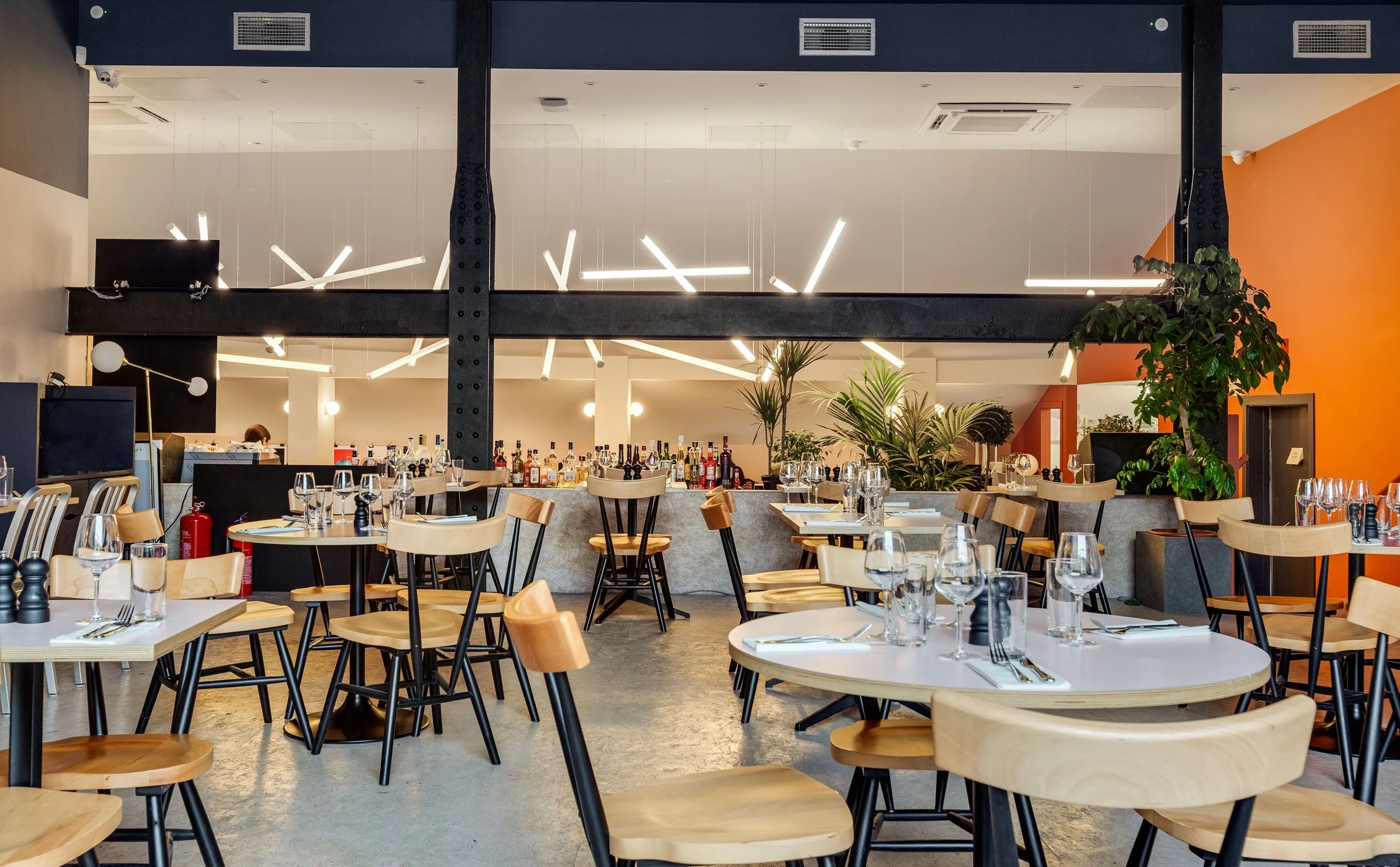 Elegant restaurant interior with wooden chairs and tables set with wine glasses and cutlery, a bar area with various alcohol bottles, modern hanging light fixtures, and a large orange accent wall.