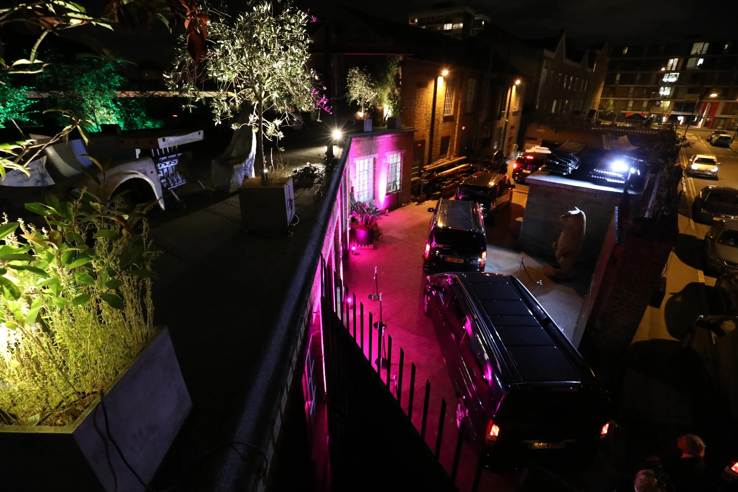 Nighttime outdoor urban scene with parked cars, lit plants, and colorful pink and white lights on a building wall.