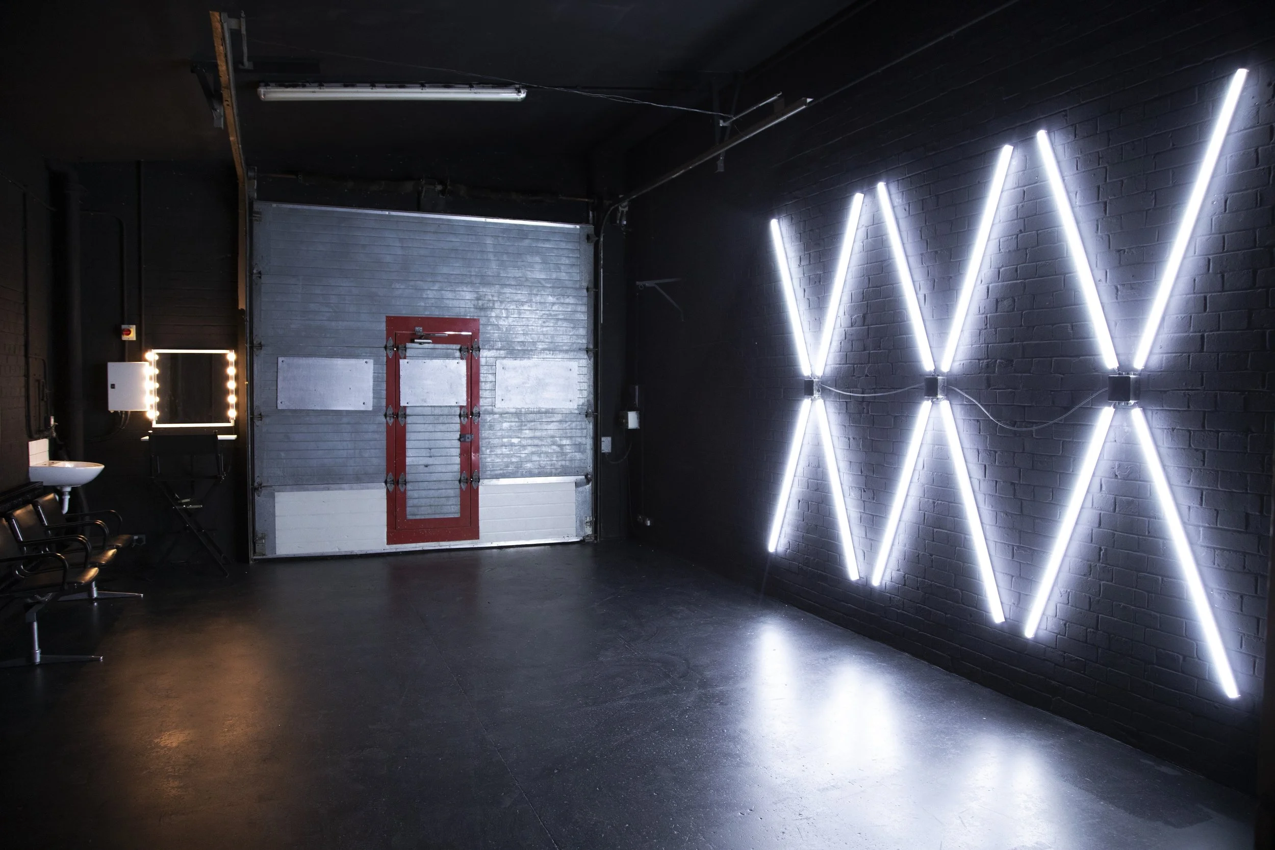 Interior of a modern salon with black walls, illuminated wall art consisting of multiple white neon lights arranged in a geometric pattern, a metal garage door with red trim, and a small sink and mirror with lights on the left side.