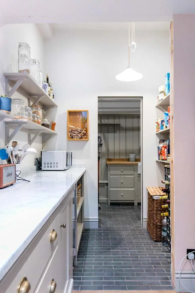 A small, organized pantry with open shelves holding jars, boxes, and canned goods, a white countertop with kitchen appliances, and a gray room with drawers and a basket at the back.