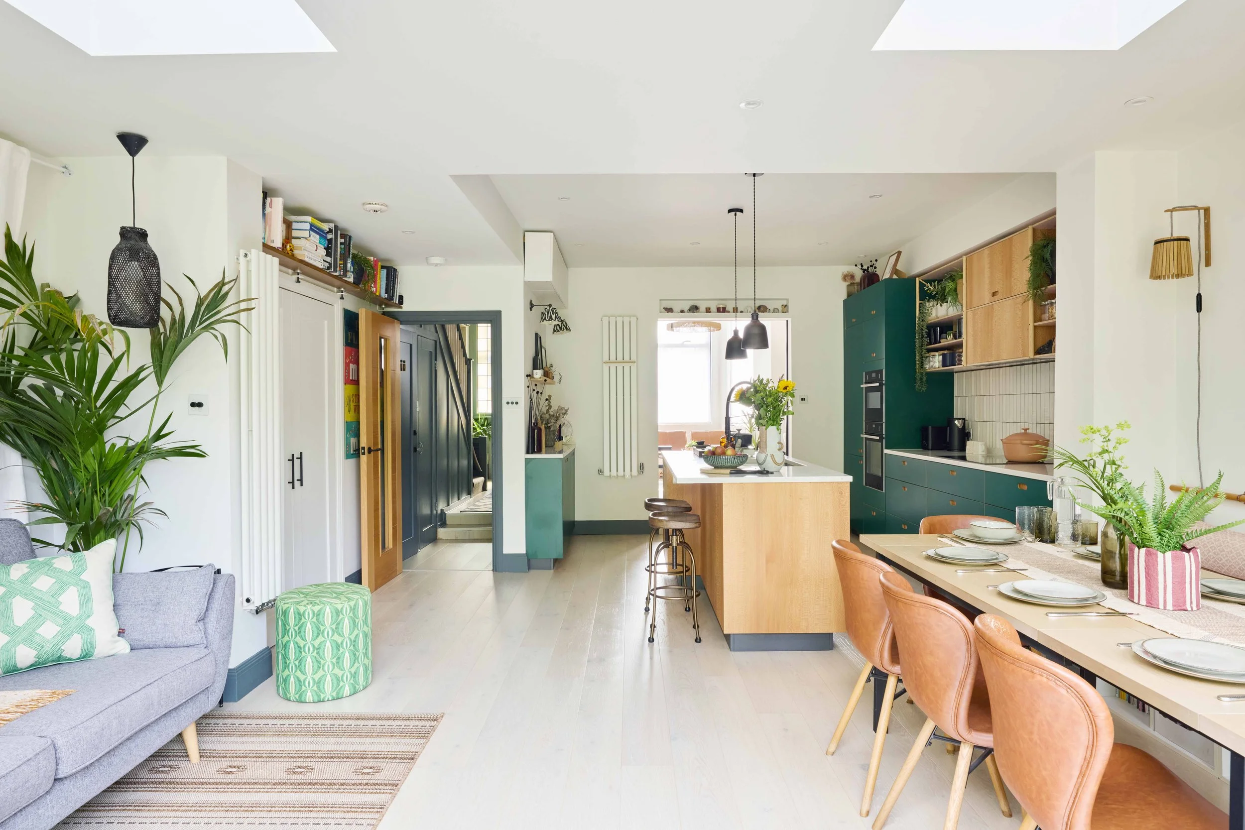 Open-plan kitchen and dining area with a green and wood kitchen, a dining table set with plates and glasses, and a living area with a gray sofa, plants, and decorative pillows.