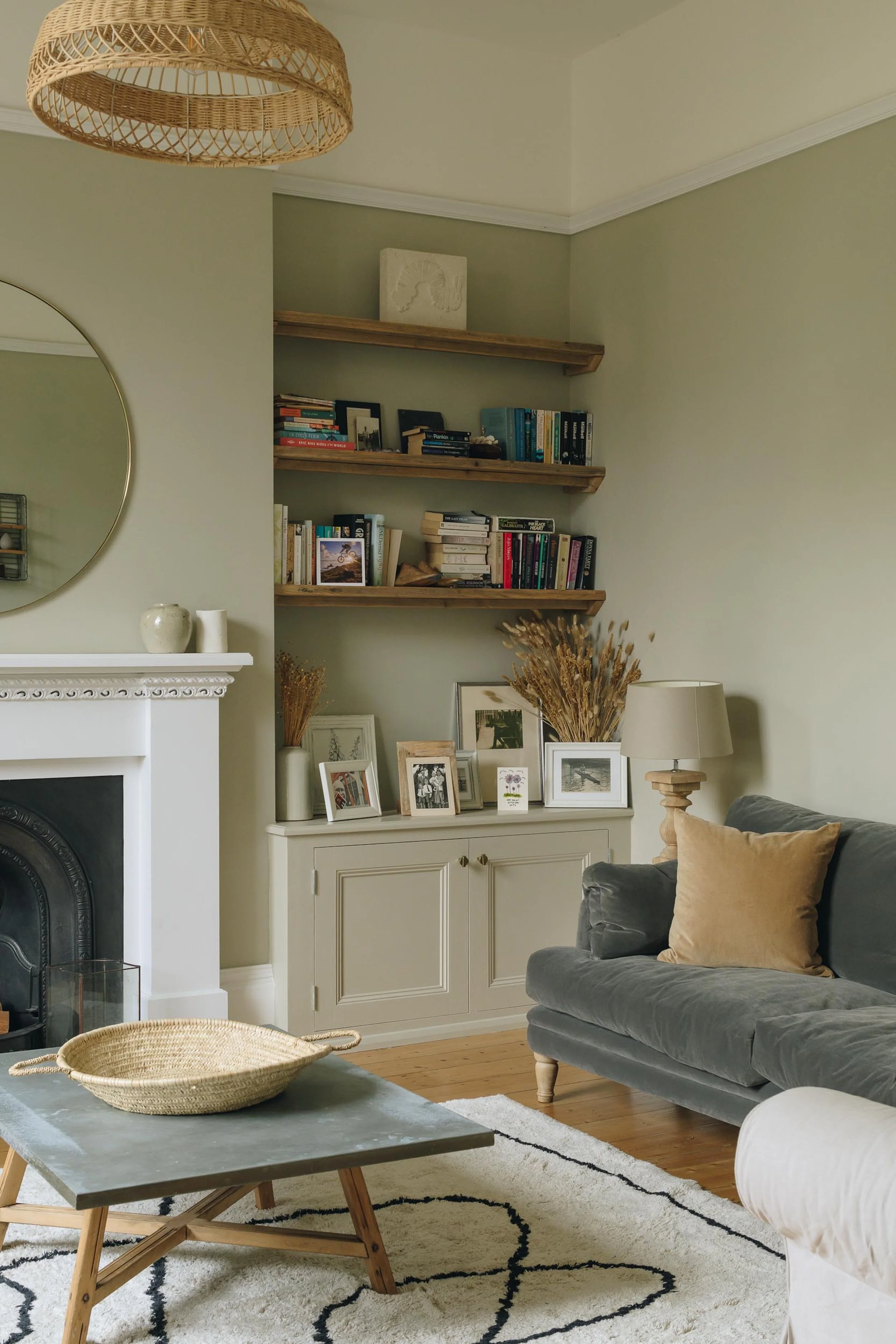 Living room with a green wall, a white fireplace, a gray sofa with a tan pillow, a black and white patterned rug, a small wooden coffee table with a woven basket, and a white cabinet with picture frames and dried plants. Three wooden shelves with boo