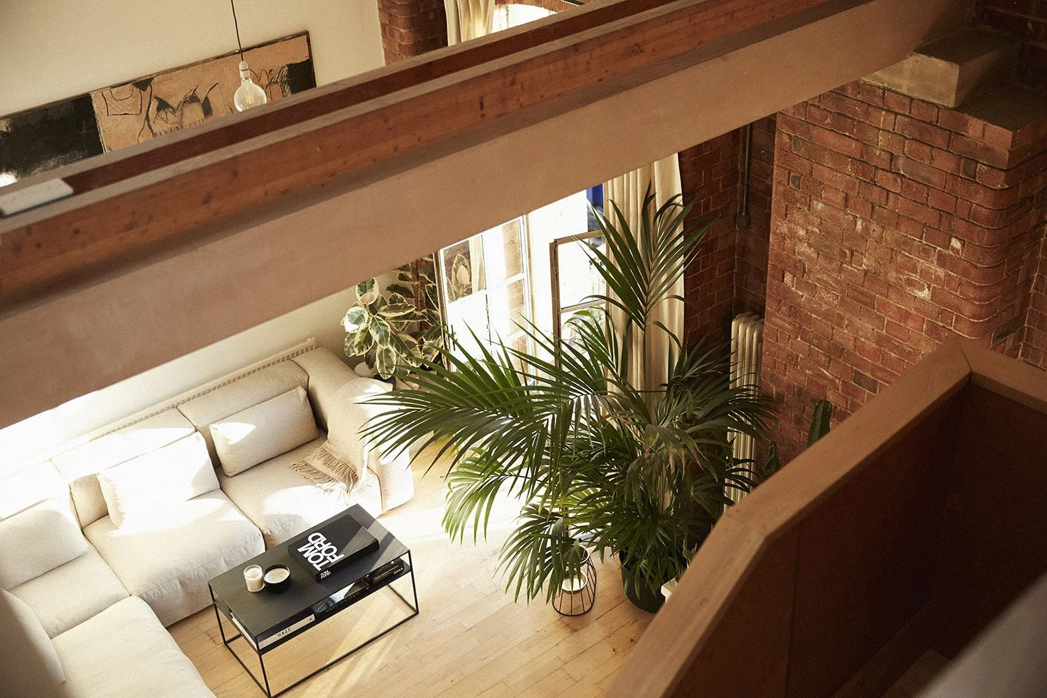 View of a cozy living room from above, featuring a cream-colored sectional sofa, a black coffee table with candles and a magazine, large green plants, windows with curtains, and exposed brick walls.