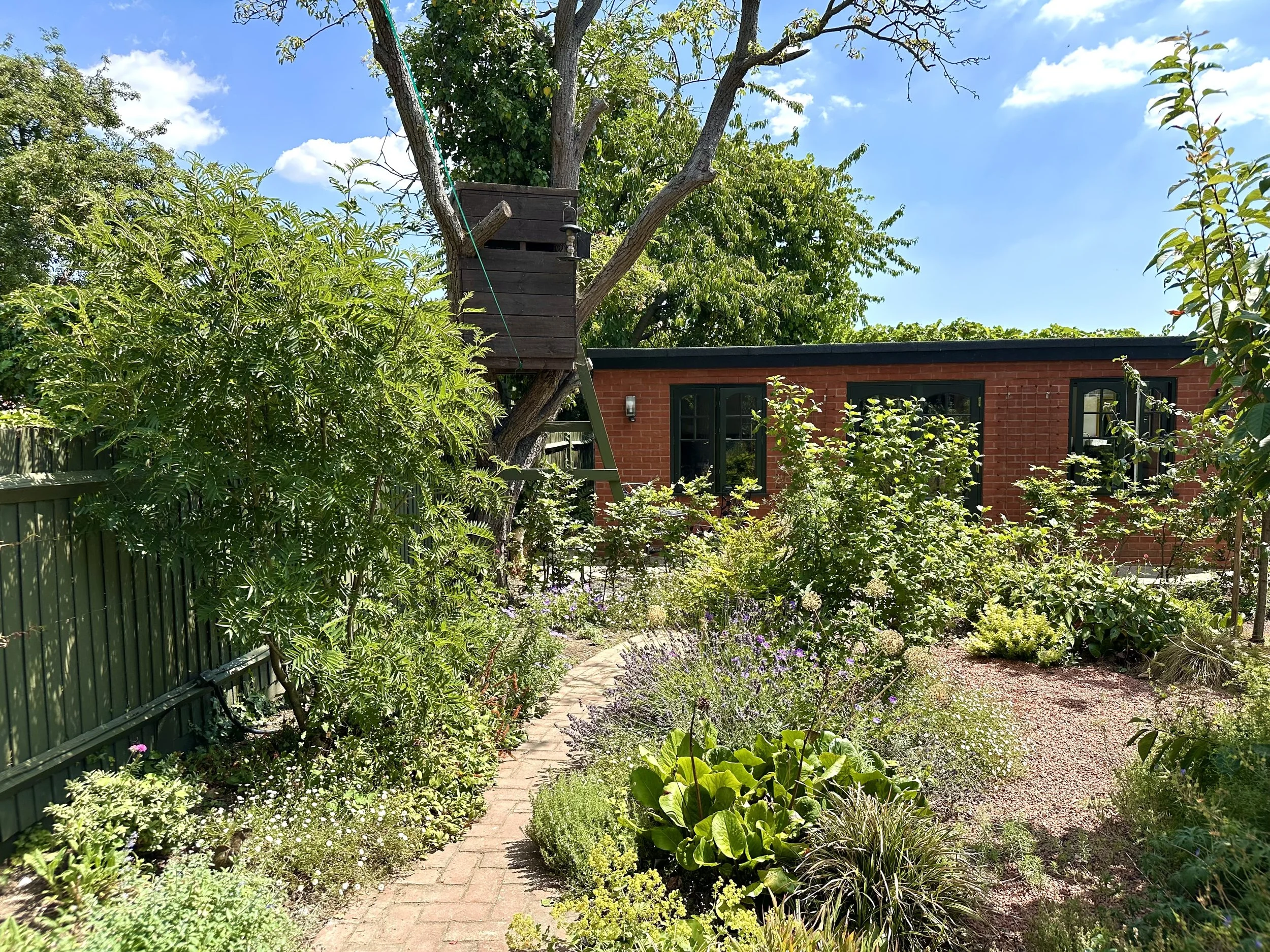 A backyard garden with a brick house in the background, surrounded by lush greenery, trees, and various flowering plants. A small brick pathway weaves through the garden.