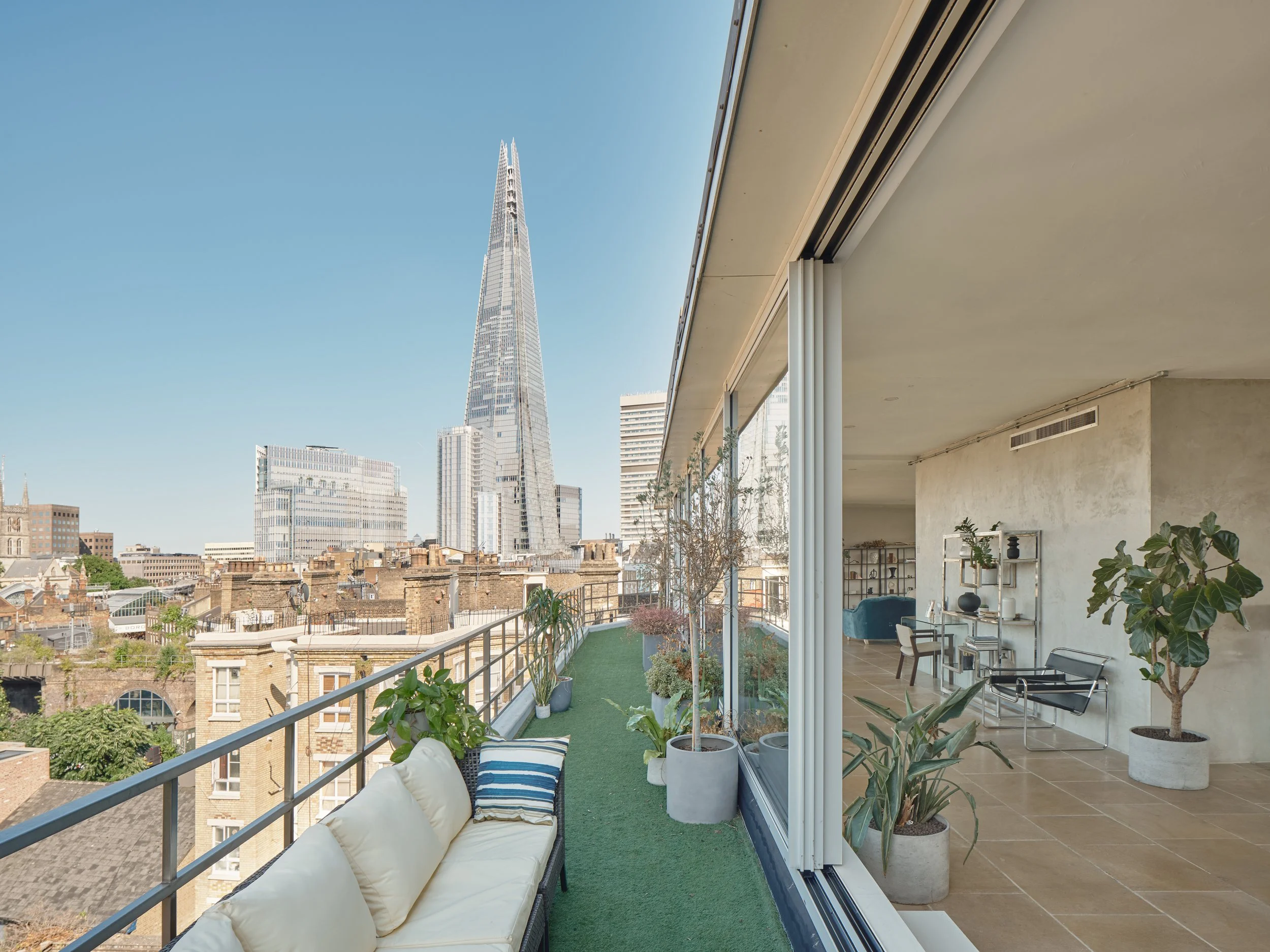 The image shows a rooftop terrace with outdoor seating, potted plants, and a view of the cityscape, including the Shard building in London.