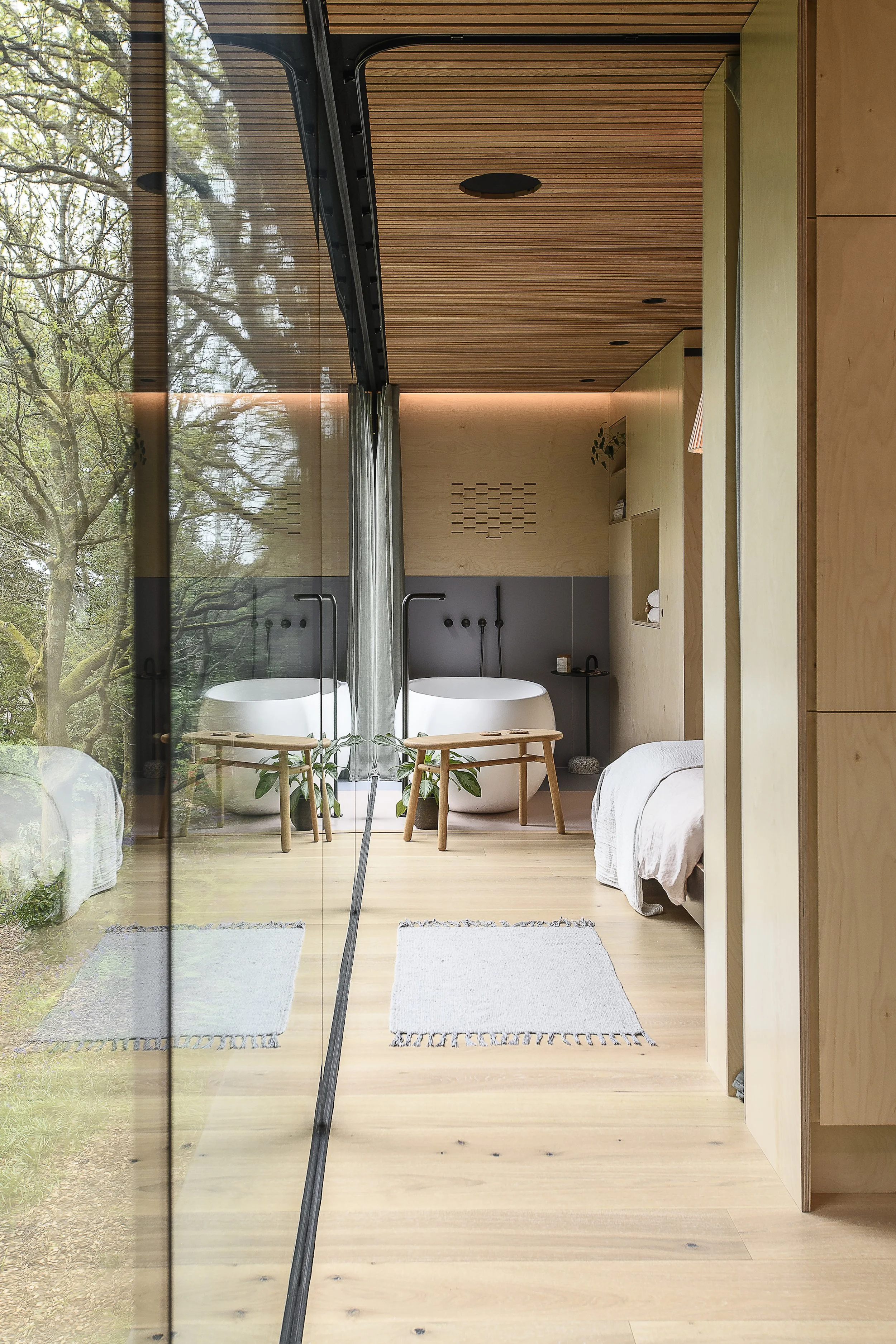 A modern bedroom with a wooden ceiling and floor, featuring a glass wall reflecting trees outside, two white freestanding bathtubs with black fixtures, small benches, and a bed with white linens.