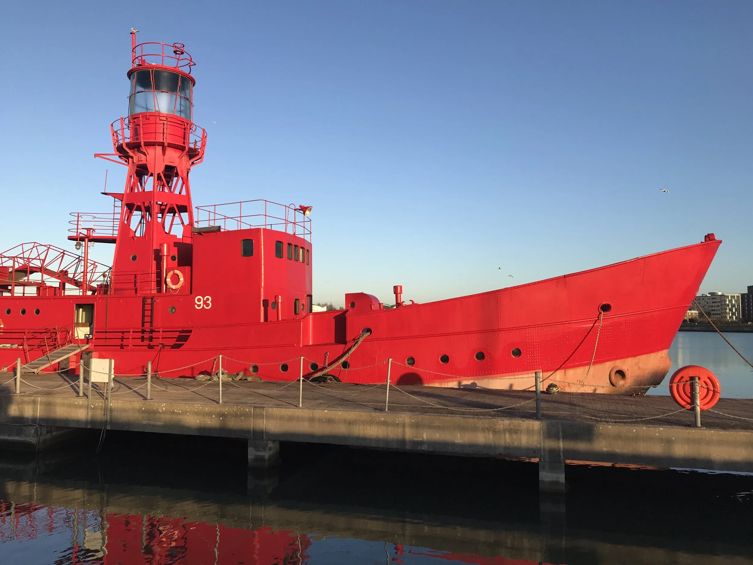 Red lighthouse ship docked at a pier, with calm water and city buildings in the background.