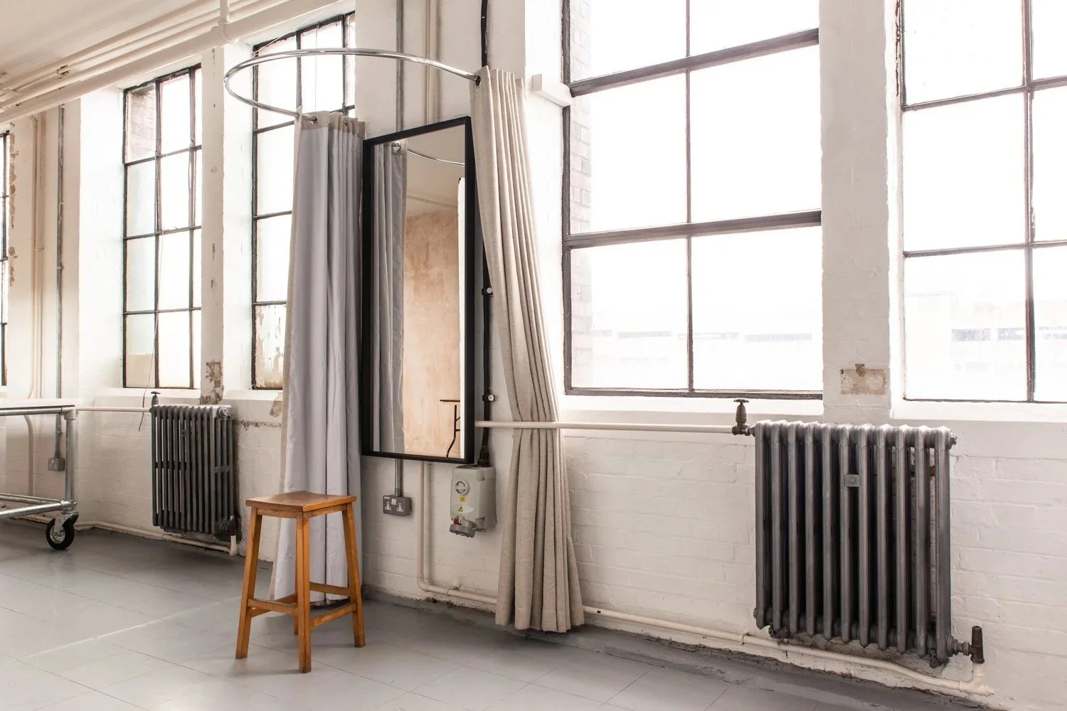 Bright industrial-style room with large windows, white brick walls, black radiators, a wooden stool, a mirror, and beige curtains.