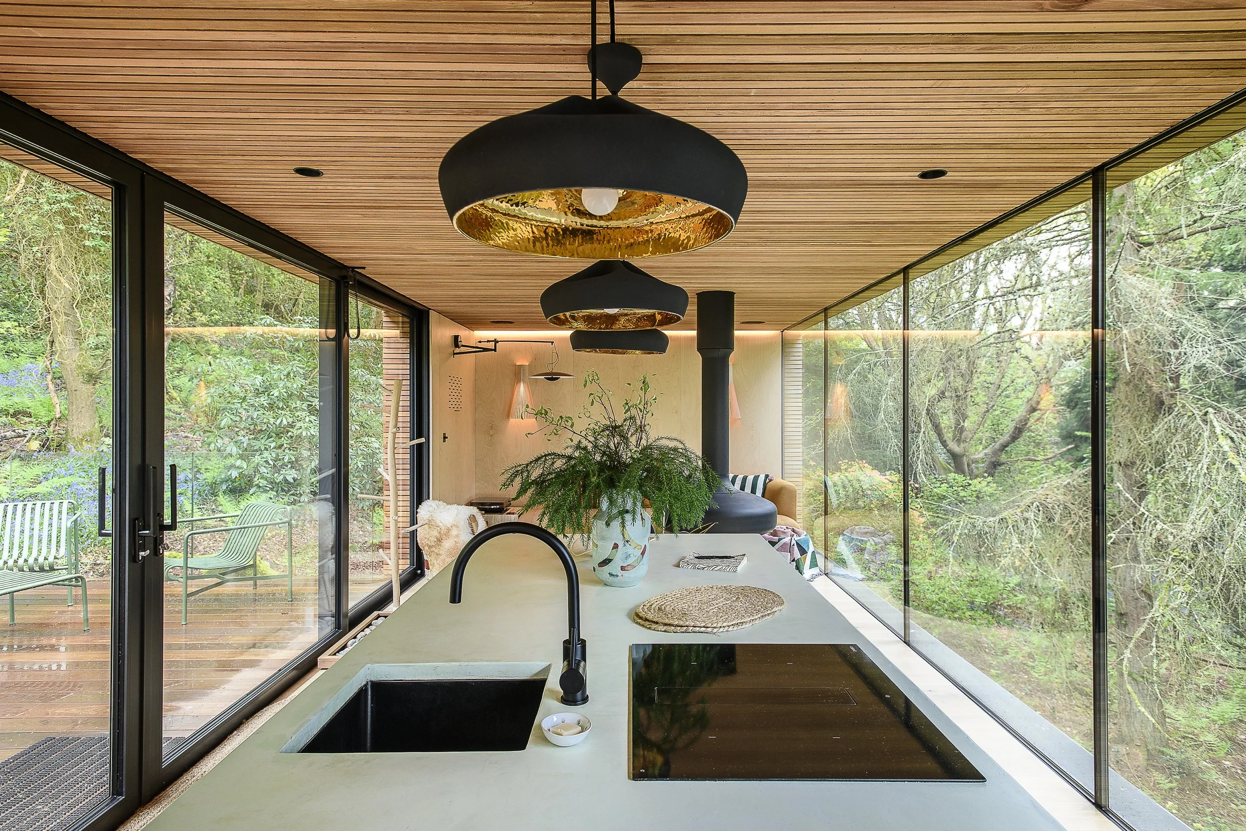 Modern kitchen with a light-colored countertop, black sink and stovetop, large glass walls revealing a lush outdoor garden, hanging black pendant lights with gold interiors, a potted plant on the counter, and a wood-paneled ceiling.