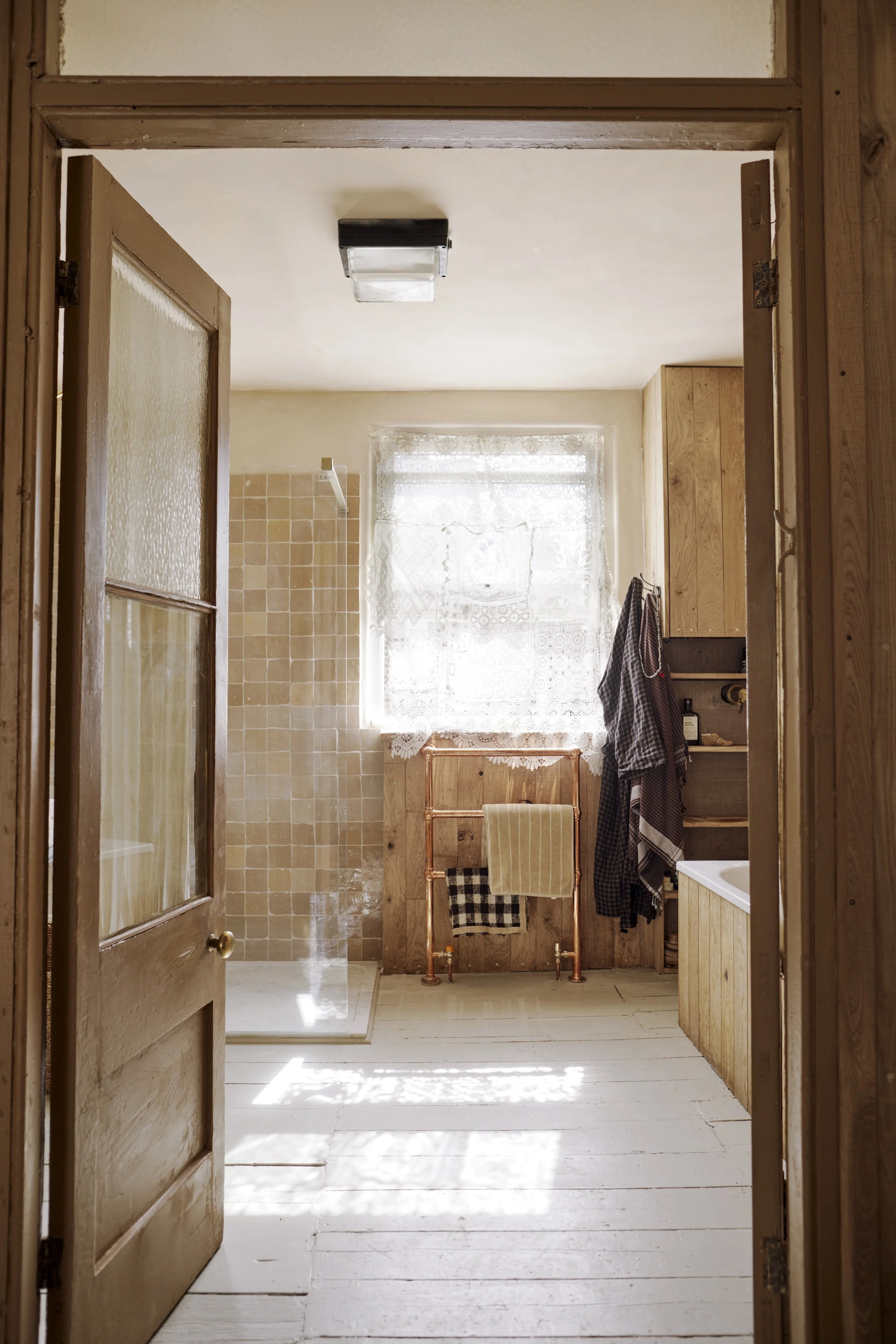 A rustic bathroom with wooden walls, a window with lace curtains, a shower with tan tiles, and a copper towel rack with towels hanging.