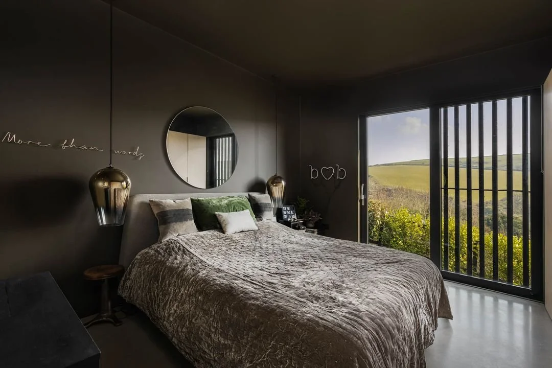 Modern bedroom with dark walls, circular mirror above the bed, gold hanging pendant lights, and a large window with vertical bars overlooking a landscape of green rolling hills.