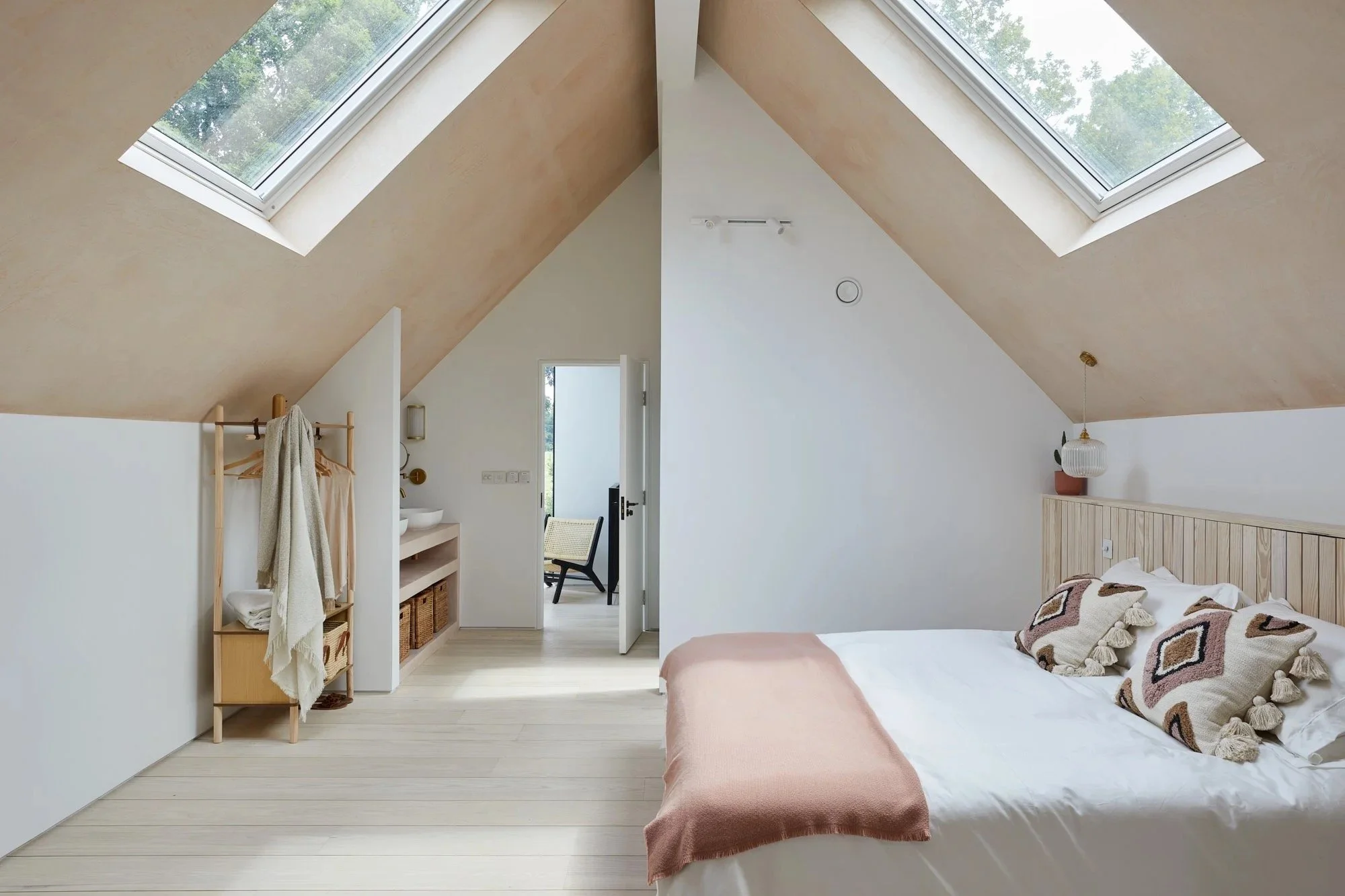 A bedroom with a bed, cozy pillows, and a pink blanket, under a sloped ceiling with skylight windows, a small open door leading to a balcony, and minimal decor.