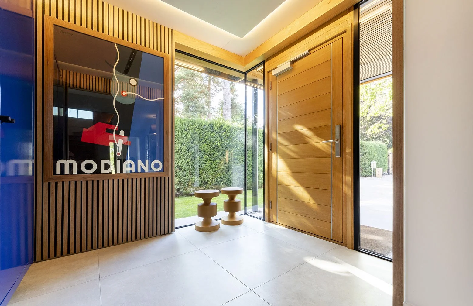 Entryway with wooden door, glass side panels, two small wooden stools, and a framed poster with a stylized house and the word "MODIANO" inside a modern home.