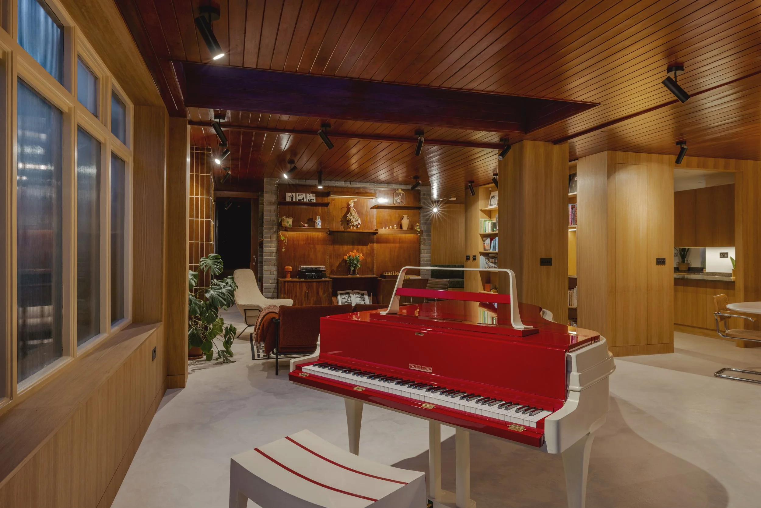 Interior of a room with a red and white grand piano, wooden walls, shelves with books and decor, a seating area with chairs and plants, and a wooden ceiling with track lighting.