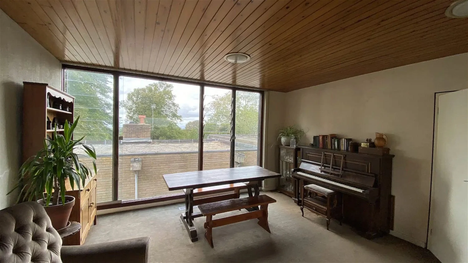 Living room with large window, wooden ceiling, piano, bookshelf, potted plant, and dining table.