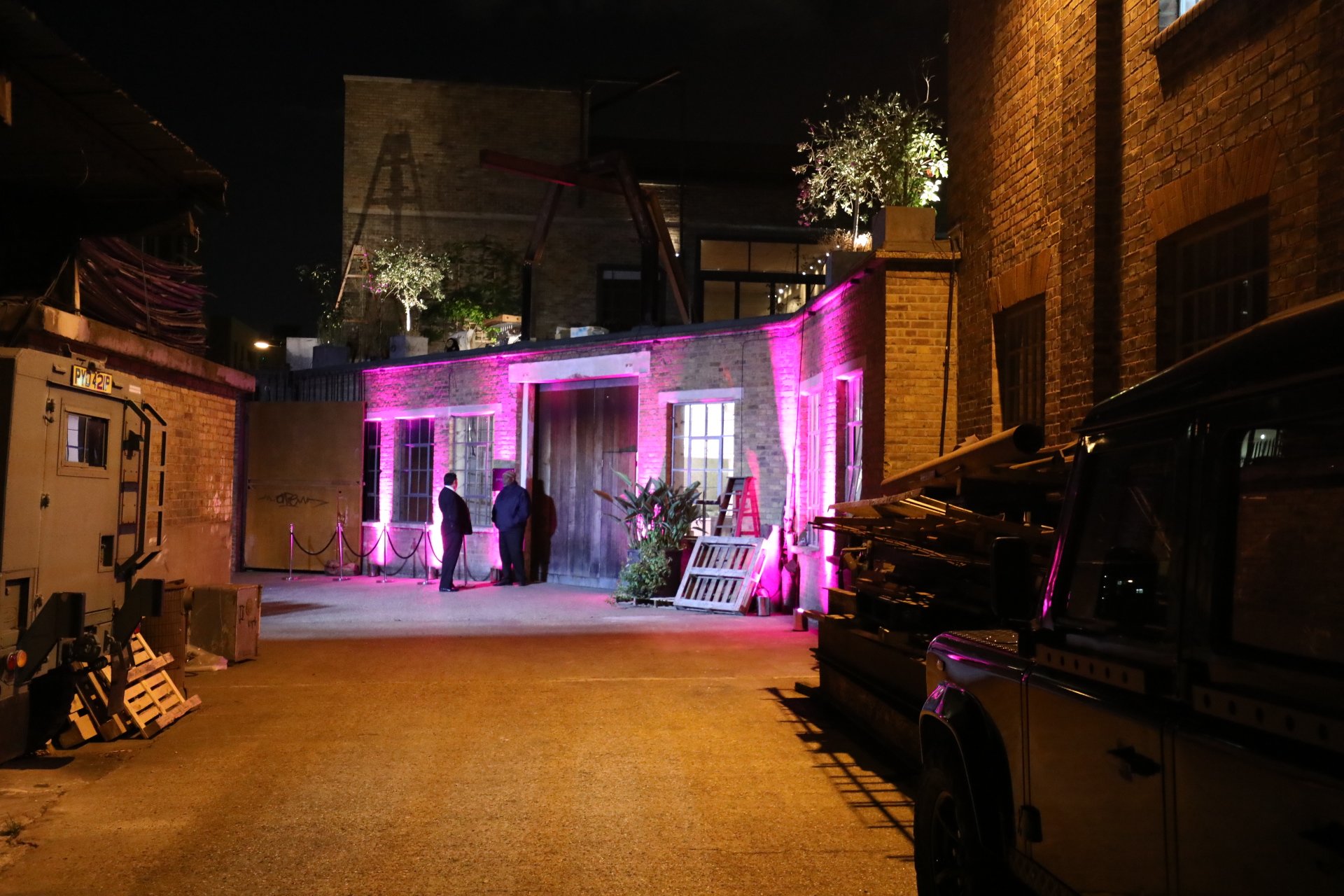 Night scene of an urban alley with a brick building illuminated by pink and purple lights, three people standing and conversing near the building, with construction materials and a vehicle nearby.