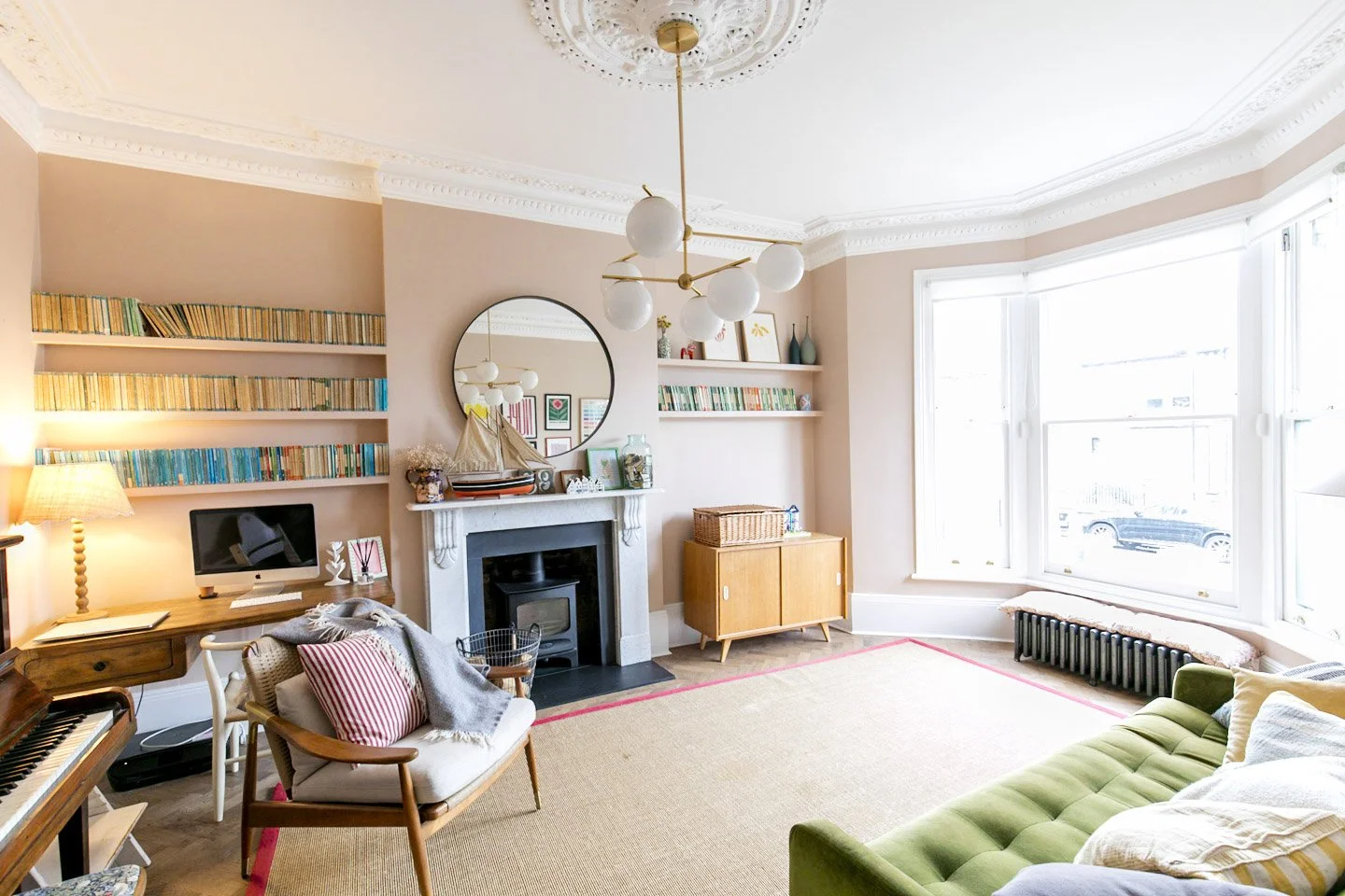 A living room with a white fireplace, round mirror, bookshelf, green sofa, and large bay window.