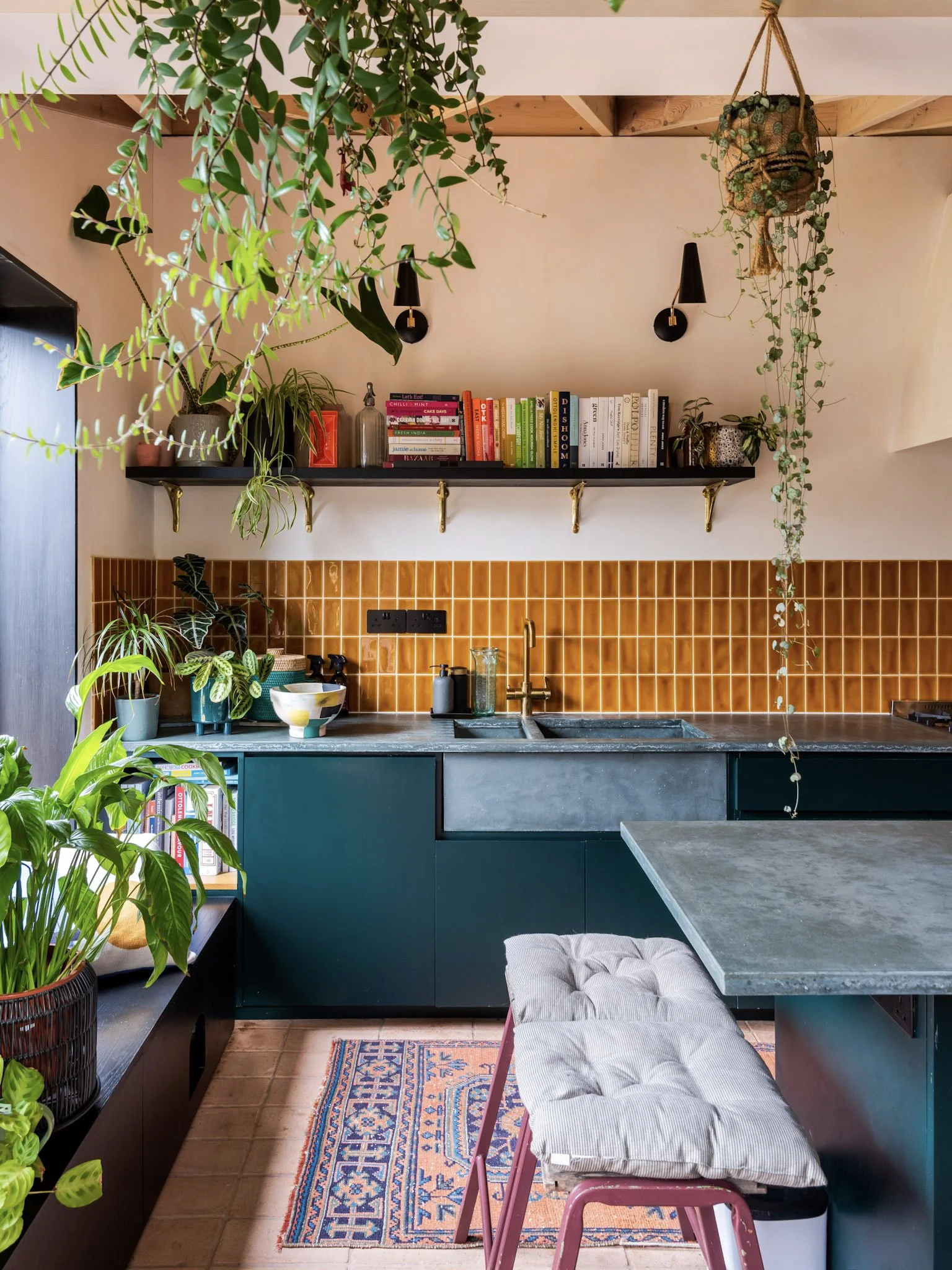 A cozy kitchen with green cabinetry, an orange tiled backsplash, and a concrete countertop. It features hanging plants, open shelves with books, and a small dining area with a cushioned bench and a patterned rug.