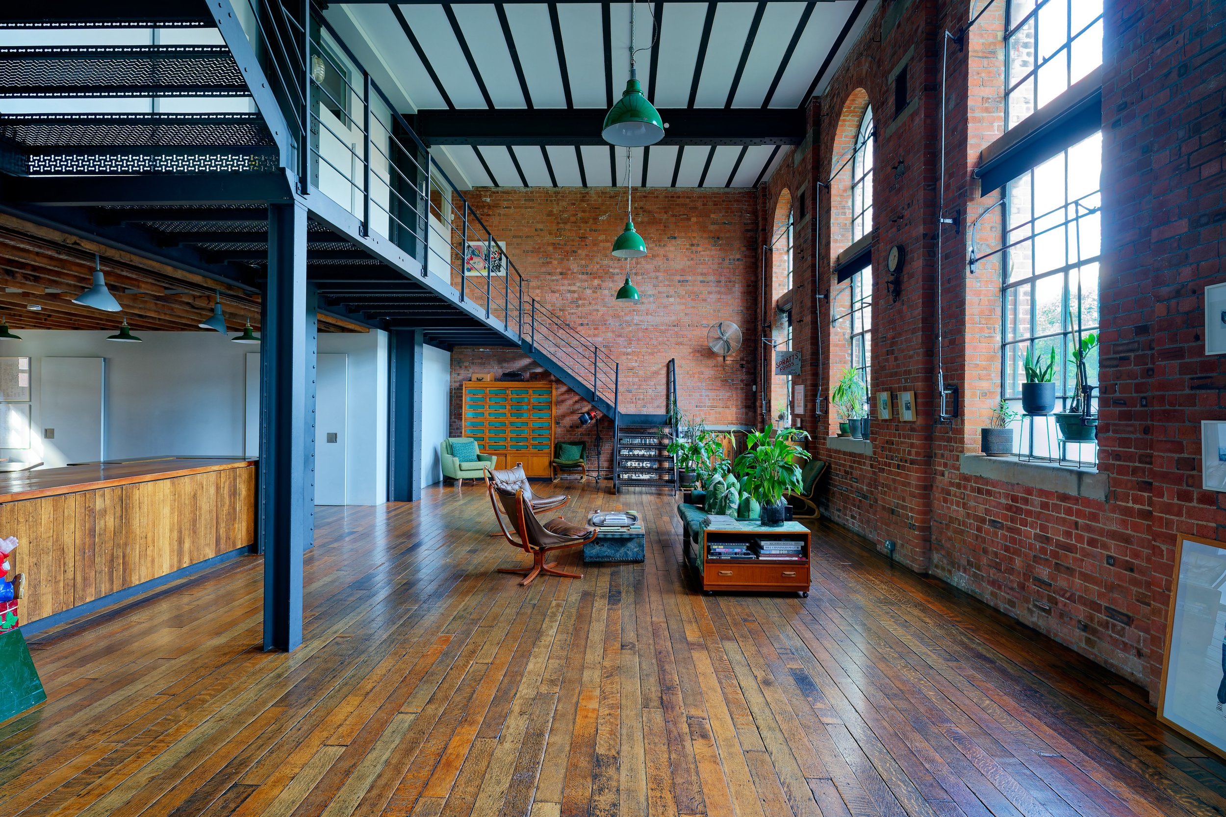 Spacious loft with brick walls, large windows, wooden floors, and eclectic furniture, including chairs, chairs, plants, and wall decor.