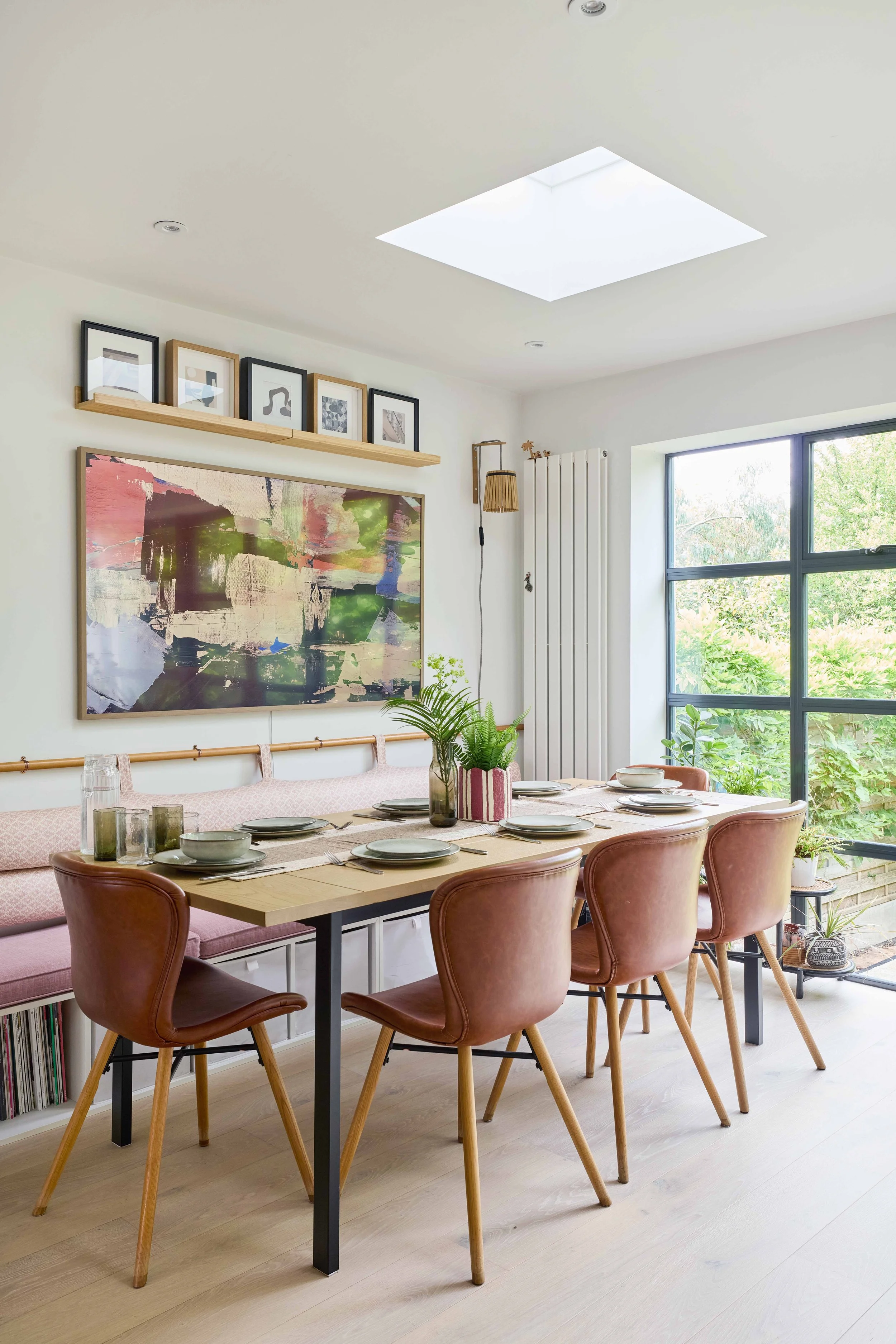 A dining room with a wooden table set for six, pink upholstered chairs, a large window with greenery outside, framed artwork and photographs on the wall, and a skylight overhead.