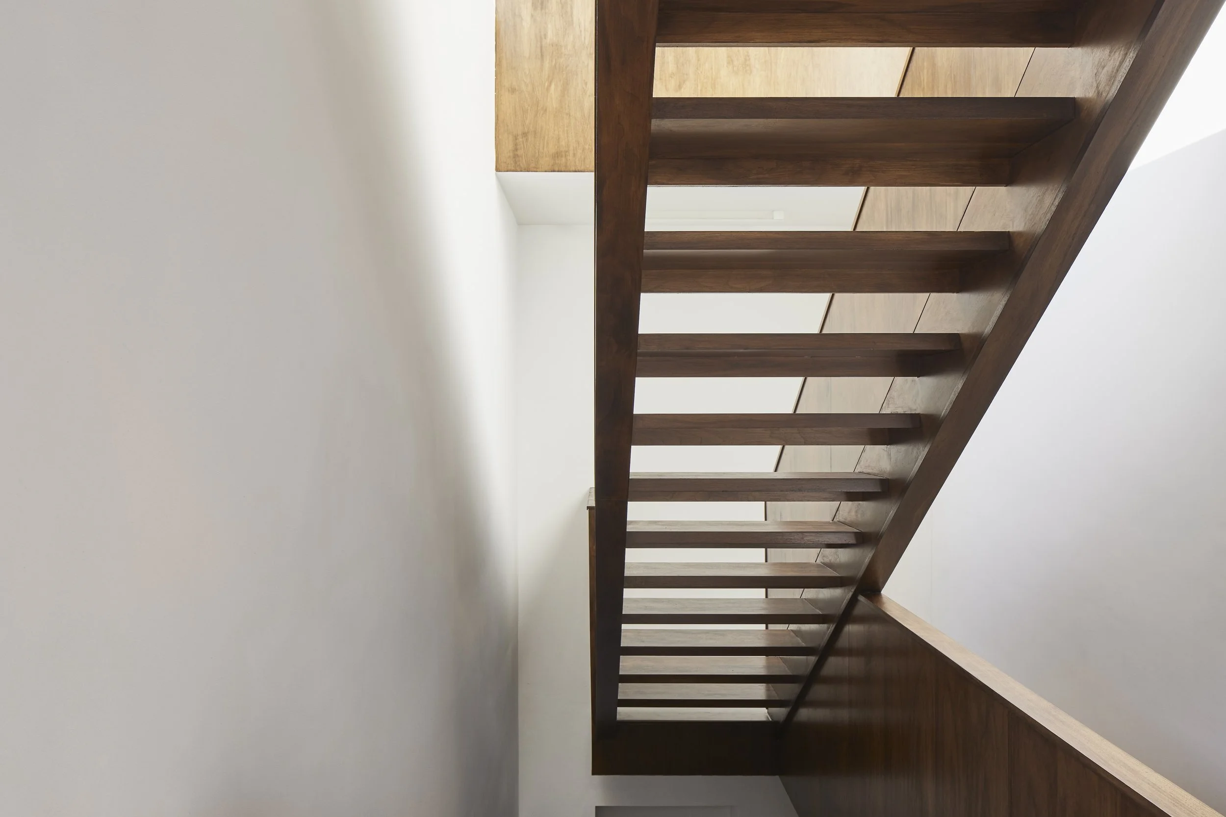 Photo of a modern staircase with wooden railing and steps, taken from a low angle against a plain white wall.