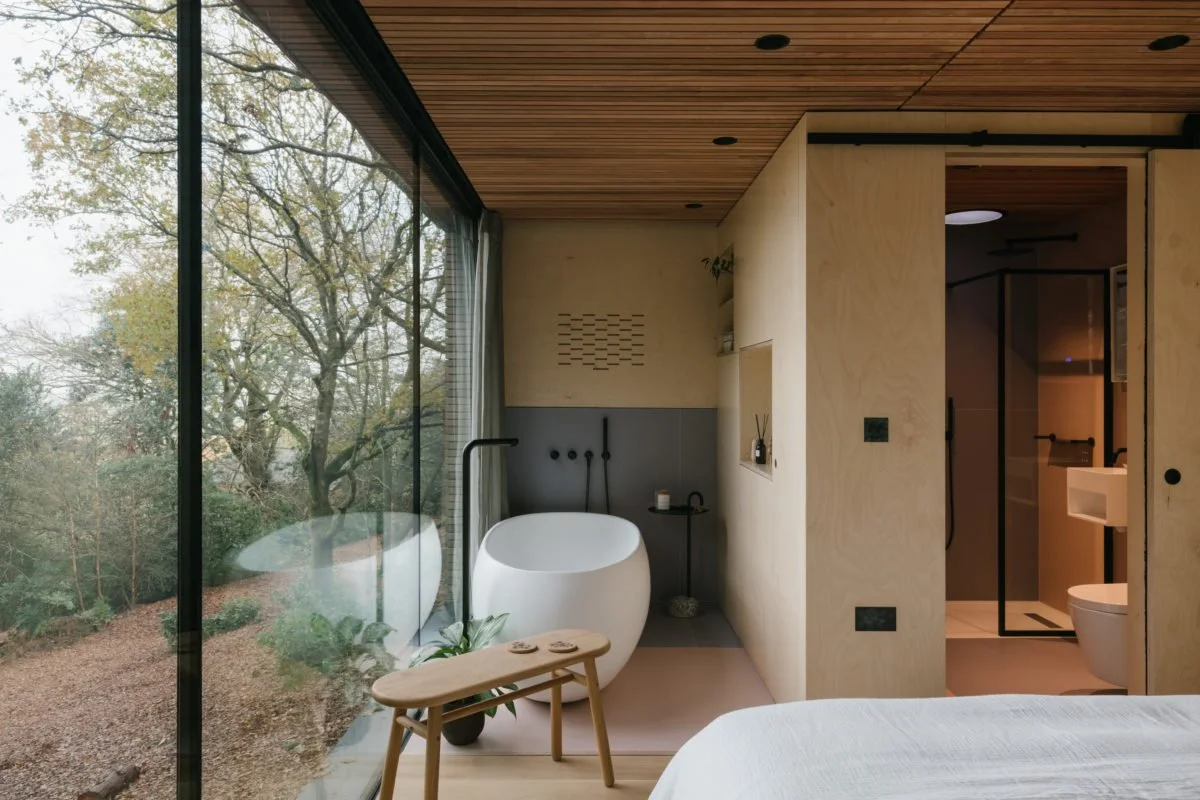 Modern bathroom with a freestanding bathtub near large glass windows overlooking trees, with wood-paneled ceiling and minimalist decor.