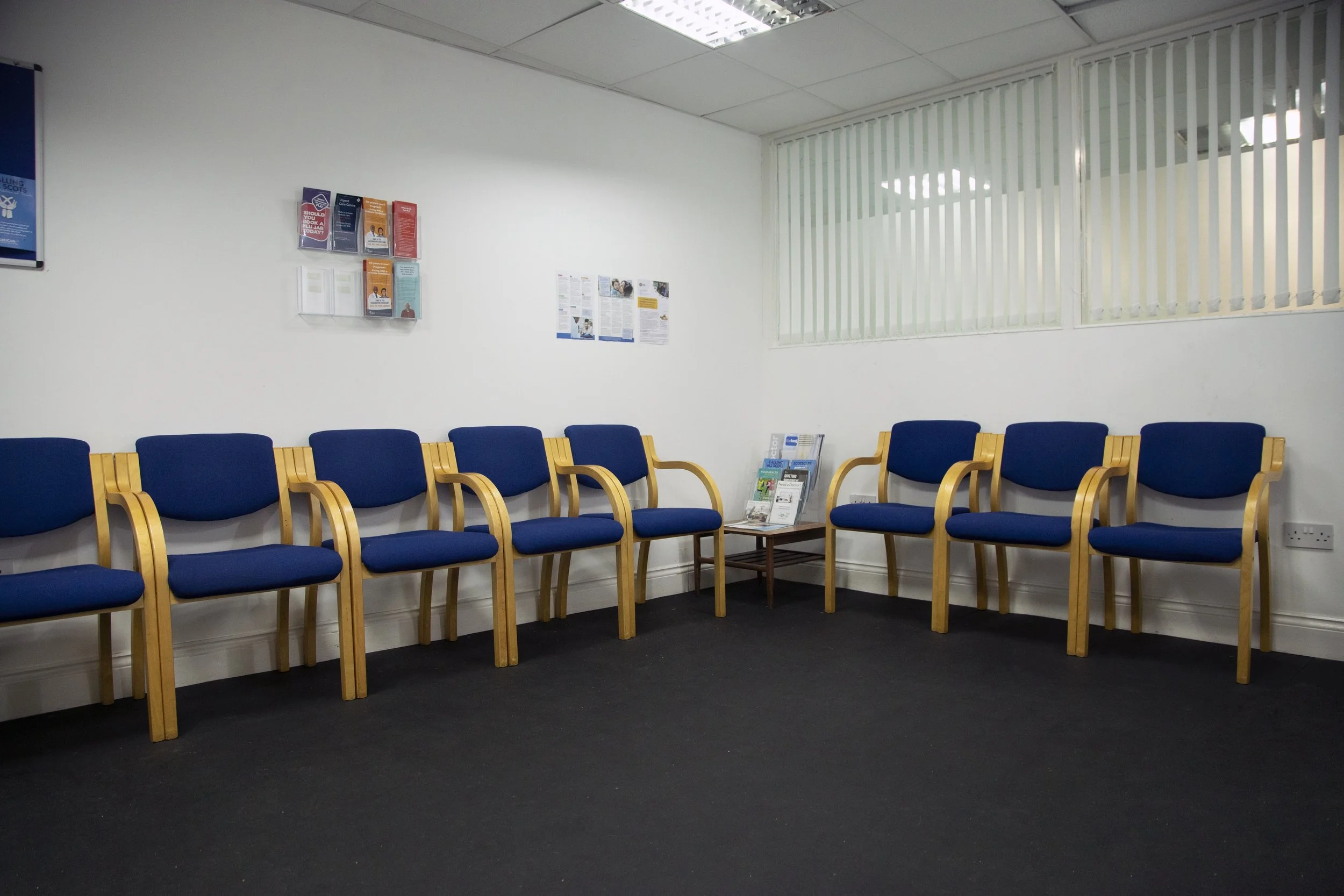 Empty waiting room with blue chairs, a small table with magazines, white walls, and vertical blinds on the window.