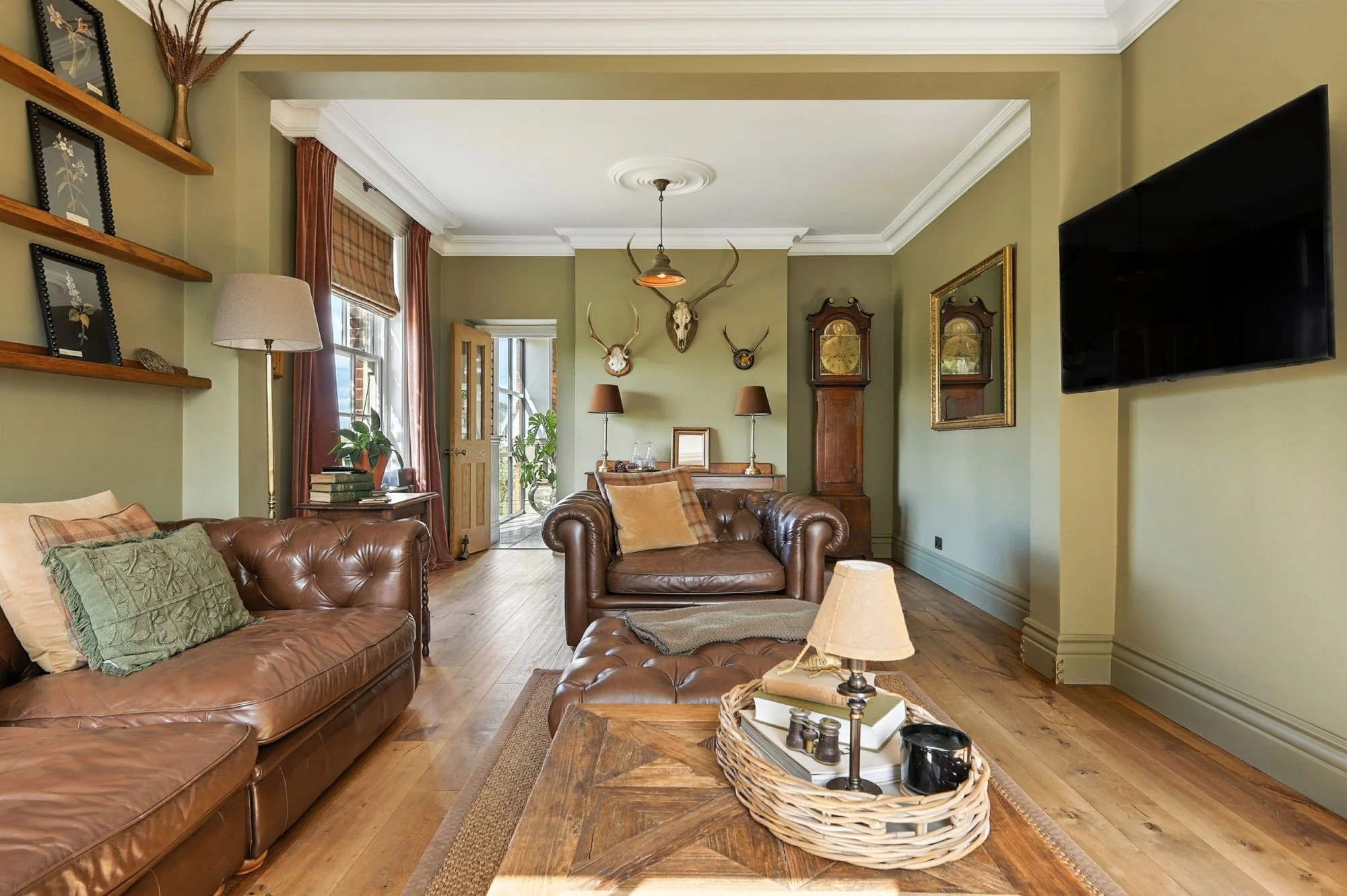 Living room featuring two brown leather sofas, wooden coffee table with decorative items in a wicker tray, wall-mounted flat-screen TV, vintage wooden grandfather clock, artwork, and mounted deer and animal skulls with antlers on the walls.