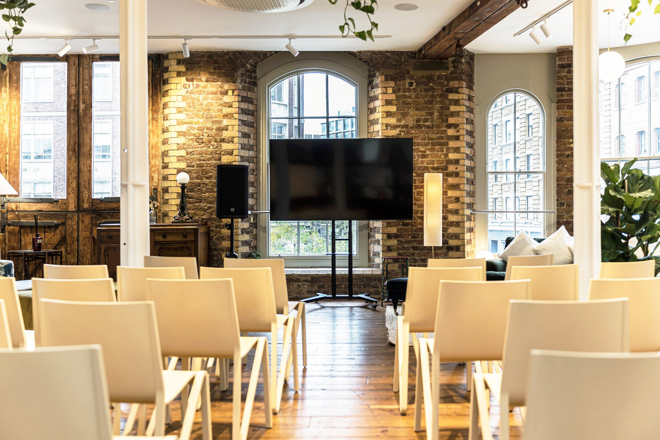 An indoor event space with rows of beige chairs facing a large flat screen TV mounted in front of a brick wall with arched windows. The room features exposed brick, wooden floors, and tall windows letting in natural light.