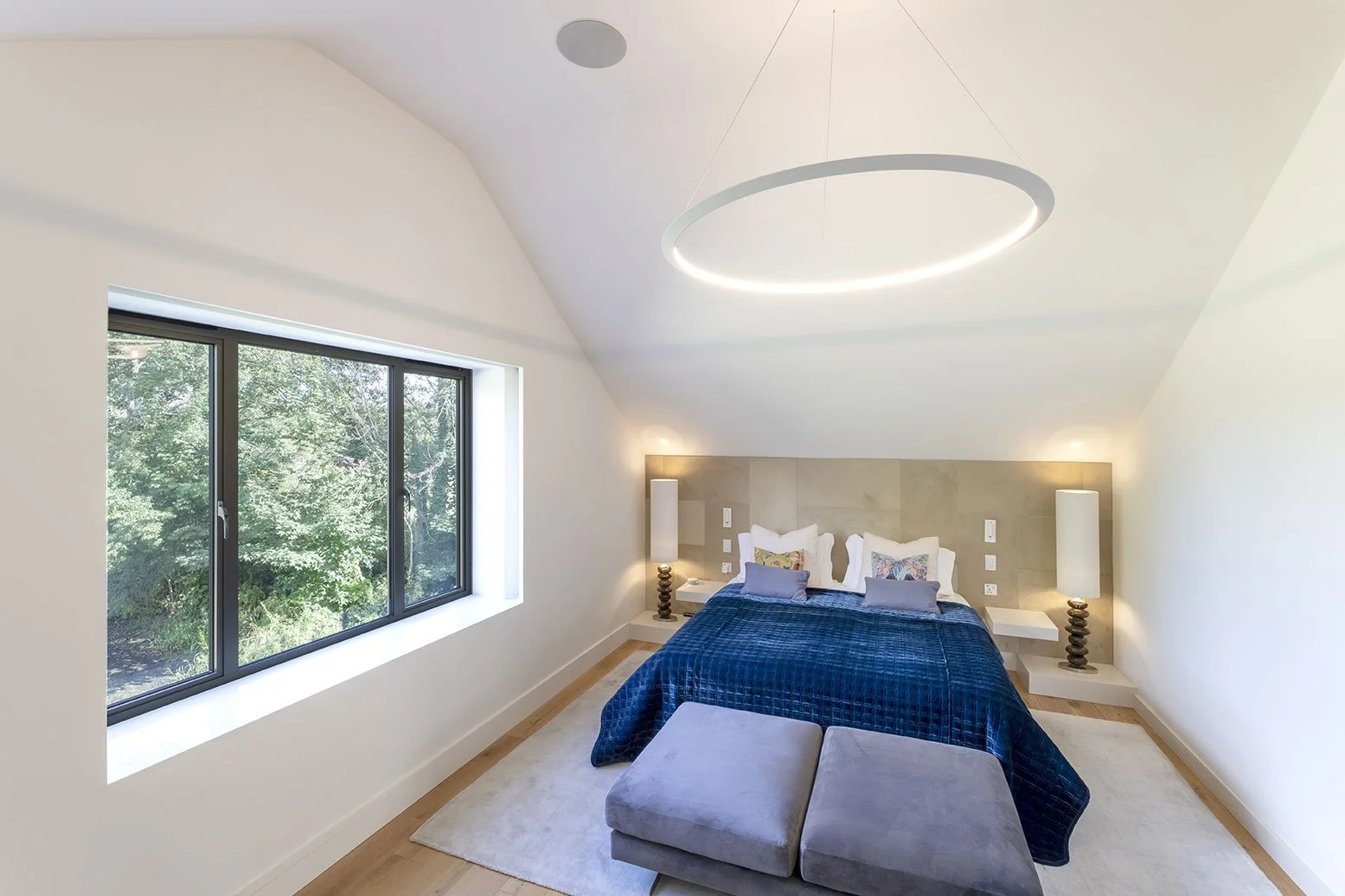 Modern bedroom with large window, cream walls, beige headboard, dark blue quilt, accent pillows, two white floating bedside tables with lamps, circular ceiling light, and gray ottomans at the foot of the bed.