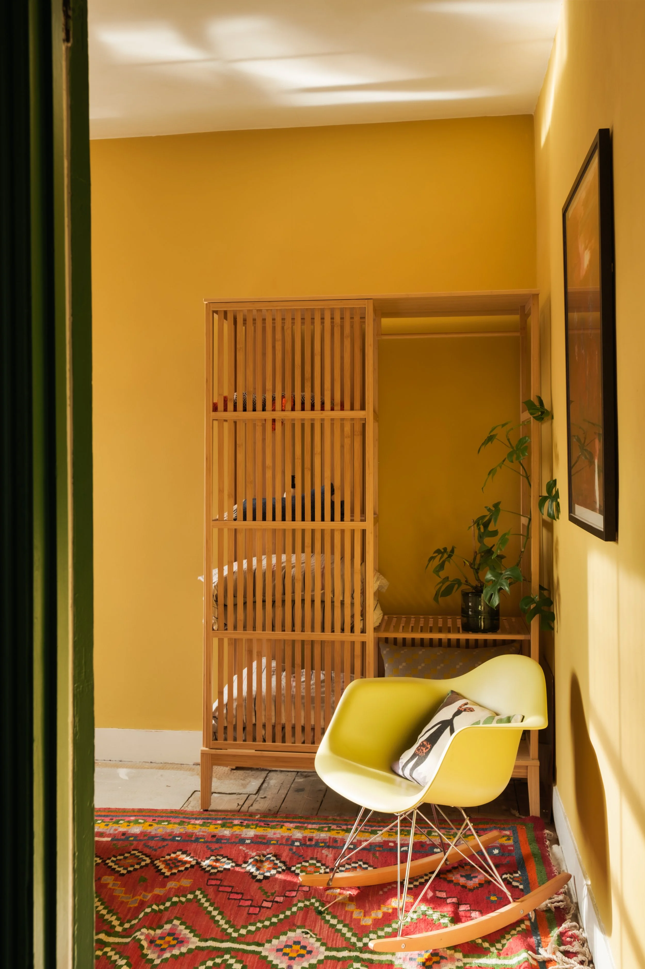 View of a cozy corner with a yellow wall, a modern yellow rocking chair with a decorative pillow, a colorful patterned rug, a wooden storage unit with open slats, and a framed picture on the wall, with a potted plant nearby.