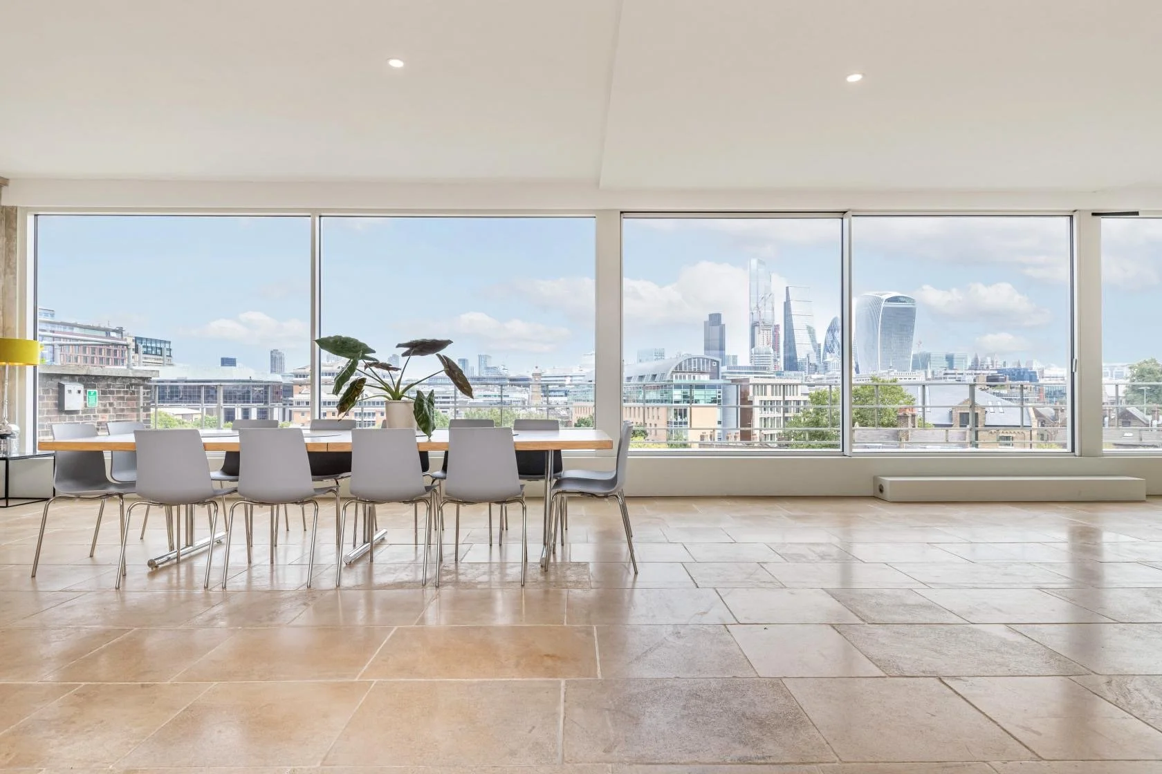 Modern office conference room with a long wooden table, white chairs, a potted plant in the center, large windows showing a city skyline, and tiled flooring.