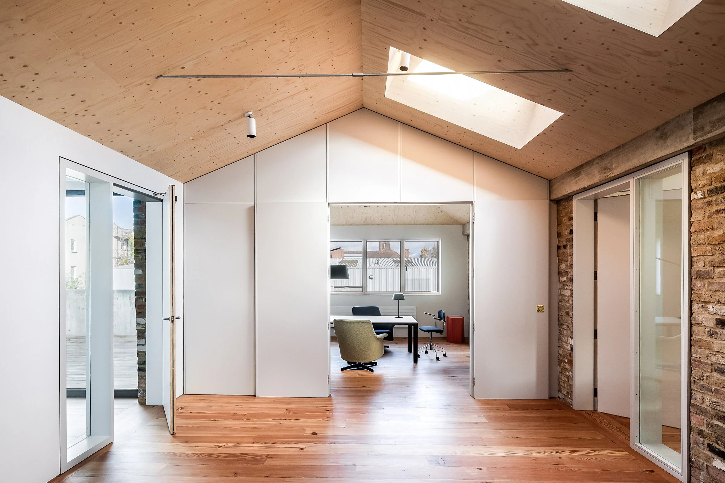 An interior view of a modern room featuring wooden flooring, white walls, a vaulted ceiling with a skylight, and an office area with a desk, chairs, and large windows.