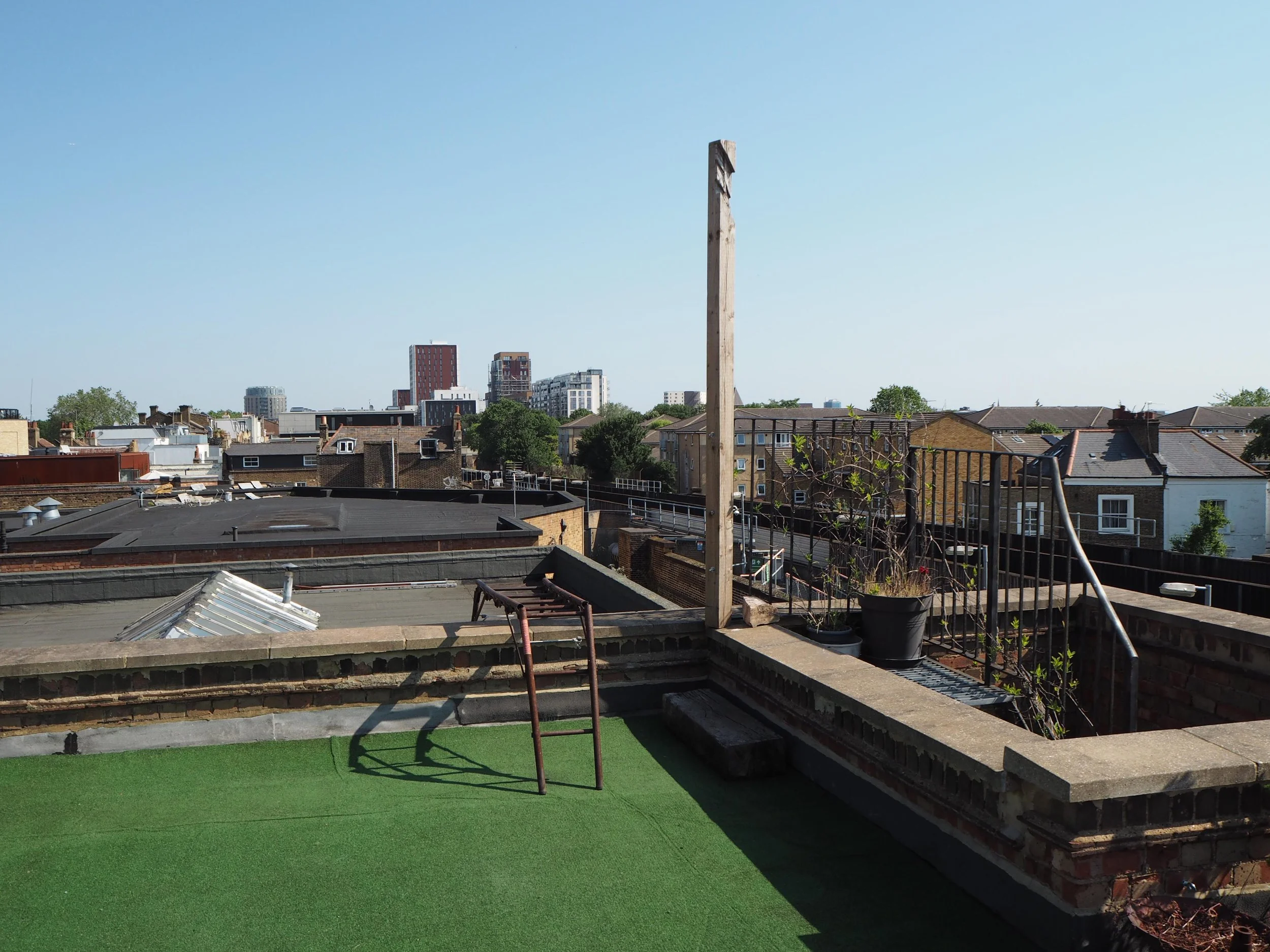 Rooftop view of a cityscape with buildings, trees, and a clear blue sky, featuring a small rooftop garden with potted plants and a metal railing.